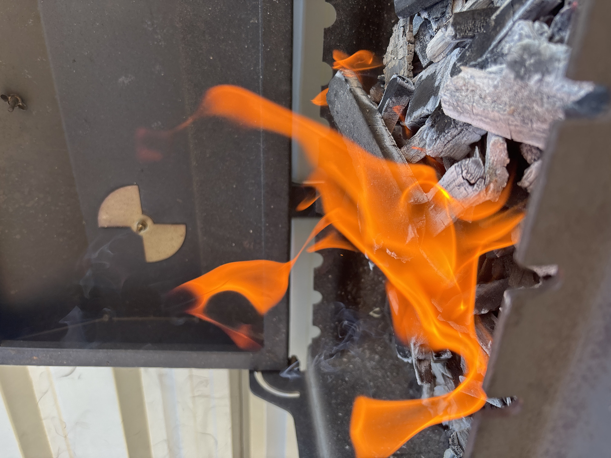 A close-up shot captures a metal barbecue grill in use. Flames are visible, consuming charcoal briquettes. The grill features a vent with a nuclear symbol. Smoke is emanating from the burning charcoal. The environment suggests an outdoor setting, but the location is not identifiable as Chaikovsky, Russia from the visual data.