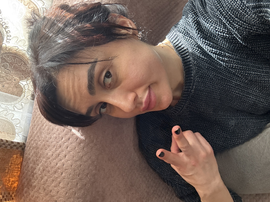 The scene, set indoors in the city of Bolnisi, Georgia, depicts a single adult female in a close-up, selfie-style shot. She has dark brown hair, a fair complexion, and dark eyes, gazing slightly to the viewer's right with a faint smile on her lips. She wears a dark charcoal-grey ribbed knit sweater, a thin silver-colored chain necklace, and a silver-colored cluster earring in her right ear. Her left hand is raised in the lower-left quadrant of the frame, forming a 'finger gun' gesture with the index and middle fingers extended; the nails on these fingers are painted dark black. The subject reclines against a textured light-brown upholstered surface, consistent with a sofa backrest or armrest, which occupies the background behind her. A light-colored patterned curtain is partially visible in the upper right background, suggesting a window or light source beyond. The environment is well-lit, presumably by natural light.