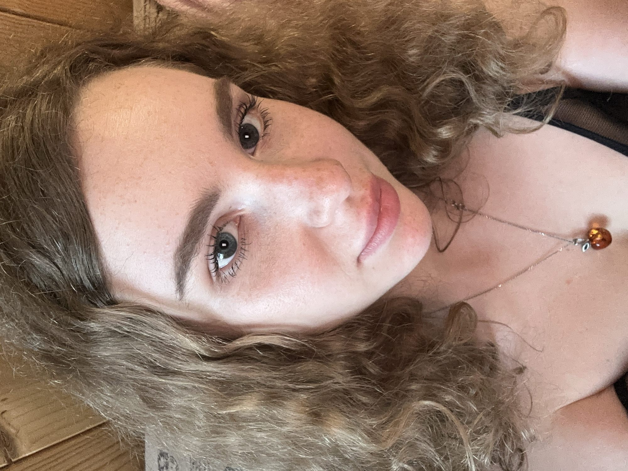 A close-up, overhead portrait features a young woman with long, curly, light brown hair. She lies on her back, looking directly at the camera with a calm, neutral expression. Her fair skin shows subtle freckles across her nose and cheeks. She has light greyish-blue eyes framed by defined eyebrows and visible eyelashes. Her lips are naturally pink and slightly parted. A delicate silver chain necklace adorns her neck, featuring a small, amber-colored pendant resting on her chest. A hint of dark fabric, possibly part of her clothing, is visible on the right side of the frame. Her wavy hair cascades around her head, forming a substantial part of the foreground and background, which subtly reveals a blurred wooden surface on the left. The lighting is soft and even, highlighting her features without harsh shadows. There is no visible text in the image.