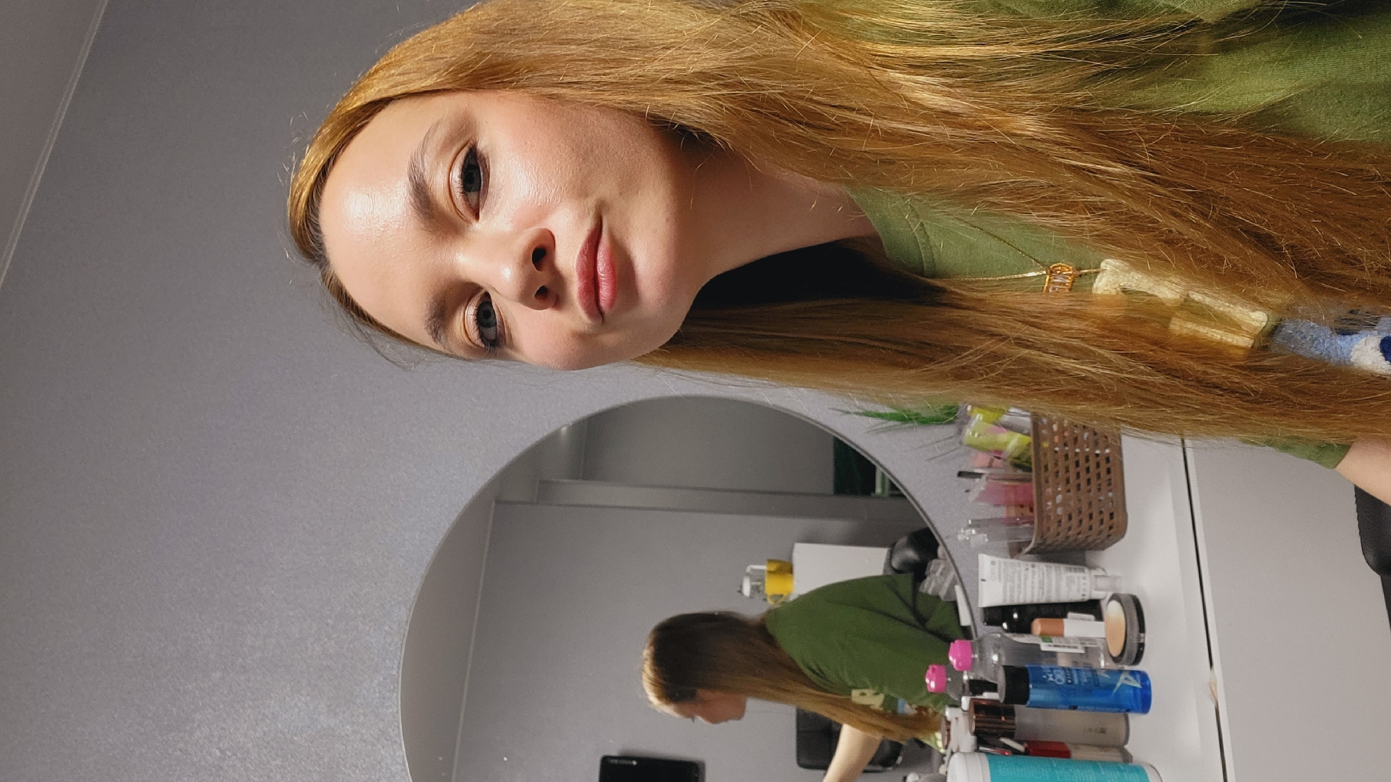 A fair-skinned female with long, auburn hair is the primary subject, situated in what appears to be a domestic setting. She is wearing a green garment, and part of a gold necklace is visible in her hair. A rounded mirror reflects a portion of the scene, including a profile view of the woman. In the mirror's reflection, a table or counter is visible with a collection of beauty products, including tubes, bottles, and containers. The wall behind the mirror is a solid, muted gray. The scene appears to be indoors, possibly a bedroom or bathroom.
Based on the location mentioned, the scene is situated in the city of Chaikovsky, Russia.