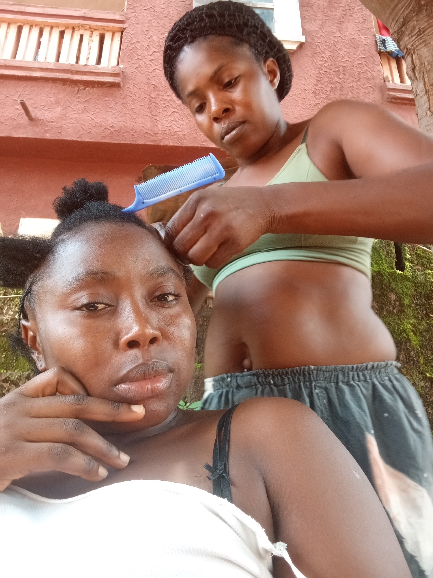 Two women are engaged in hair styling, likely outdoors, in what appears to be a residential area in Ogidi, Nigeria.
In the foreground, a woman with dark skin and natural, dark hair partially styled into a bun, looks directly at the camera with a neutral, perhaps pensive, expression. Her right hand is raised to her chin/jaw. She wears a white top.
Behind her, partially obscured, is another woman with dark skin and braided or twisted hair. She is actively working on the first woman's hair, holding a bright blue comb in her right hand and manipulating the hair with both hands. She wears a light green crop top, exposing her midriff, and dark shorts. Her gaze is focused downwards on the task at hand.
The background features a building with a distinctive reddish-orange, textured wall and a wooden balcony railing. To the far right, a large tree trunk is visible, and patches of green moss grow on a surface behind the women. The lighting suggests it is daytime. There is no visible text in the image.