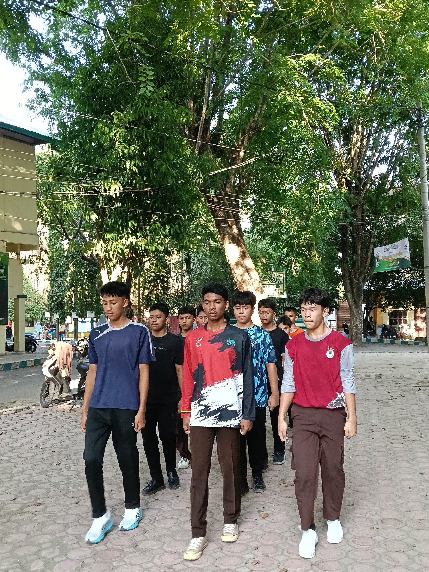 A group of approximately eight to ten young men or teenage boys is walking in a single file line on a paved path outdoors. They are dressed in casual attire, including various t-shirts (some plain, some patterned) and long pants, paired with athletic shoes. Their expressions are generally neutral and focused as they move forward.
The setting is an outdoor area characterized by abundant greenery. Large, leafy trees with dense canopies line the path, casting dappled sunlight. The path itself is made of interlocking pavers, bordered by a curb. In the background on the left, a building with a green roof is partially visible, along with a parked motorcycle and a few other people in the distance. On the right, more buildings and a utility pole are present. Overhead, numerous power lines crisscross the sky.
The time of day is bright daylight, suggesting a clear and sunny day. A banner is visible hanging between two trees on the right side, with the words "Selamat Datang" (Welcome) clearly legible, along with other Indonesian text that is indistinct. A green sign on a pole in the background on the left appears to read "UM DIN SCHOOL" or similar, hinting that the location might be a school campus in Lhokseumawe, Indonesia. The group appears to be engaged in an organized walk or activity.