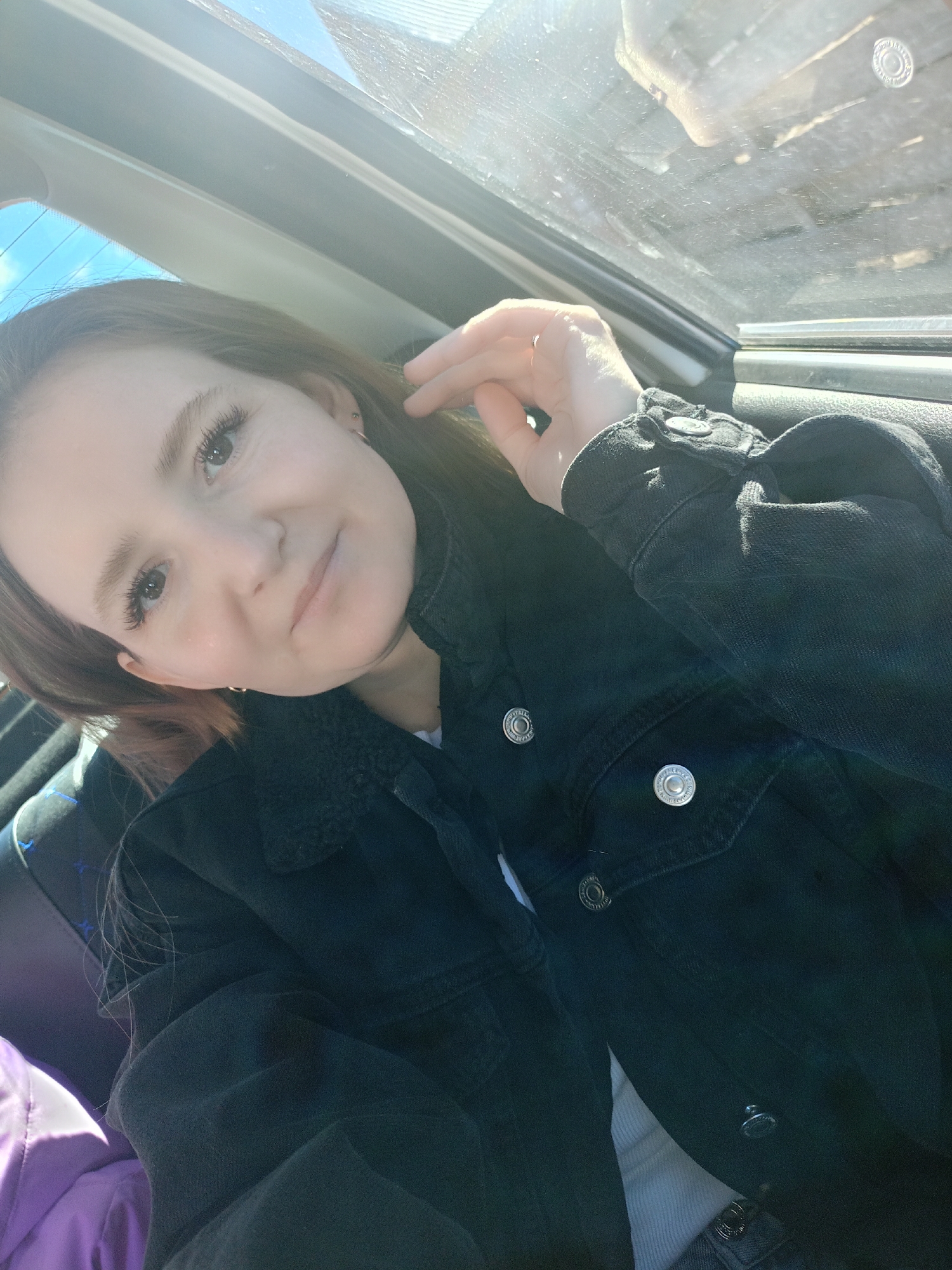 A young woman sits inside a vehicle, likely a car, due to the visible window frame and car interior components. She is wearing a black denim jacket with silver buttons and what appears to be a sherpa collar. A glimpse of a white shirt underneath the jacket is visible. Her hair is light brown, and she is holding her right hand near her ear. A portion of a purple garment is seen on the left side. The location seems to be Unknown, Unknown.