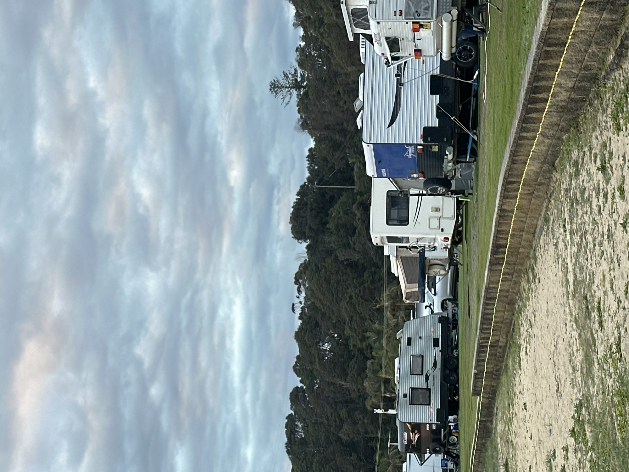 The image shows a caravan park in Nambucca Heads, Australia, with several caravans parked on a grassy area. A sandy foreground with a low stone wall runs across the bottom of the frame, separating it from the grass and caravans. Behind the caravans, a dense line of dark green trees forms a backdrop, with a cloudy, grey sky overhead. Some of the caravans are modern and well-equipped, suggesting a popular tourist destination. The overall impression is one of a serene, outdoor setting, likely near the coast given the sandy foreground.