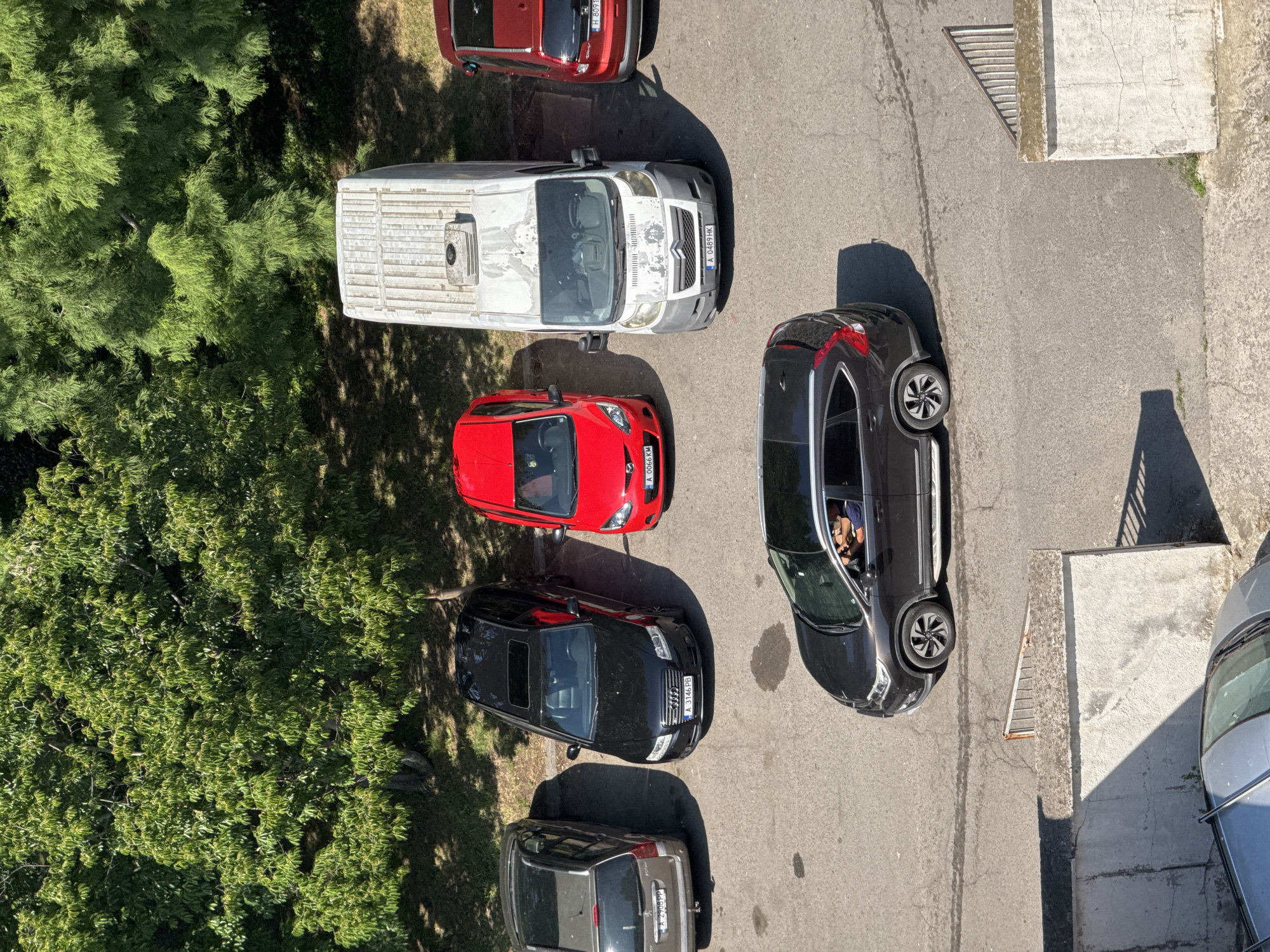 This is an aerial, eye-level shot of a parking lot with trees in the background. In the foreground, there is a tan concrete wall with a metal railing on top, and a silver car parked in front of it. Moving back, there is a dark-colored SUV with a person driving. Further back, from left to right, are a gray hatchback, a black Audi sedan, a red hatchback, a white panel van, and a red SUV. The parking lot is paved with asphalt and shows some water stains. The surrounding environment includes dense green trees casting shadows on the ground. The image appears to be taken in Burgas, Bulgaria, judging by the car license plates which feature the "A" prefix commonly used for vehicles registered in Burgas.