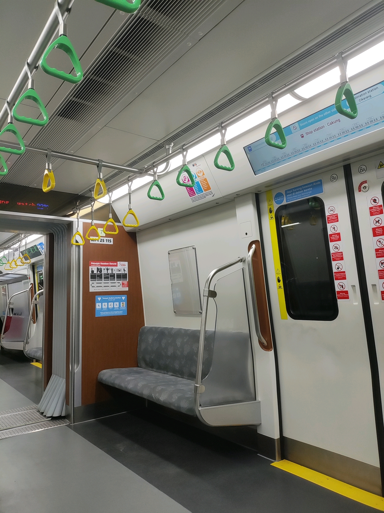 The image shows the interior of a train carriage, likely in Bekasi, Indonesia, based on the presence of Indonesian text on signage. The carriage is clean and modern, with grey flooring and white walls. Green and yellow triangular handgrips hang from metal poles along the ceiling, with some also attached to the walls. A gray bench seat with a patterned fabric is visible. To the right, a white train door with a black window and a yellow vertical stripe is present. Several warning signs with red and black graphics are affixed to the door. A brown panel with a vertical metal handrail is on the left side of the door. In the background, more handgrips and seating are visible. The overall impression is of an operational and well-maintained public transportation vehicle.