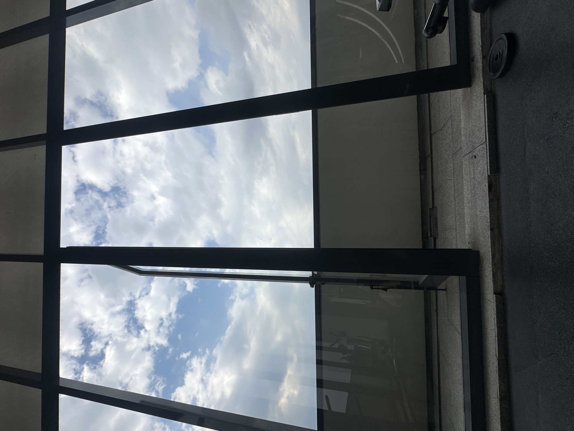 The image shows a view through a grid of black metal window frames, oriented vertically. The sky is visible through the window panes, featuring a mix of blue patches and white clouds. The window frames create a geometric pattern. To the right of the windows is a wall or column of gray concrete or stone, with some metallic exercise equipment attached. The location is indicated as Huixquilucan de Degollado, Mexico, but there are no specific markers or identifiers in the image itself to confirm this location visually. There are no people visible.