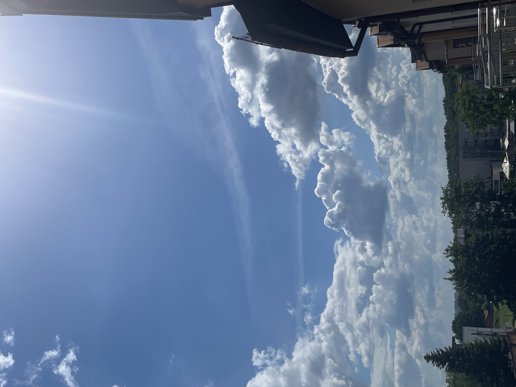 A wide-angle, vertical, eye-level shot captures a bright, blue sky filled with scattered white clouds. The sun is visible on the upper right, casting a bright lens flare. A sliver of a building's dark roofline is visible along the right edge of the frame, with a portion of its upper story showing architectural details like a chimney and metal railings. The background depicts a dense line of green trees against a hazy horizon, with some buildings partially visible behind the trees. The foreground and lower portion of the image show more of the building's exterior, including balconies with metal railings. The overall scene suggests an outdoor view from an upper floor in a city or town on a sunny day.