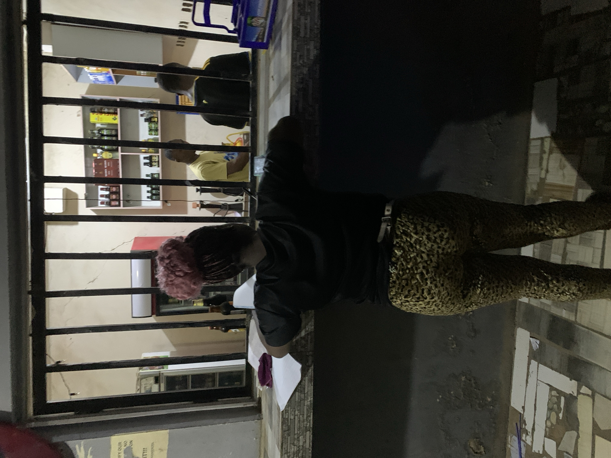 This image captures a nighttime scene in New Stadium, Nigeria, depicting a woman standing outside a commercial establishment, looking inward through a dark metal-barred opening. The woman, viewed from behind, wears a black t-shirt and leopard-print leggings, with her hair styled in an updo featuring a prominent pink/red, fluffy accessory. She leans on a tiled counter or ledge, where several papers and a dark red fabric item are visible to her left. A blue plastic crate rests on the counter to her far right. The floor in the foreground is composed of irregularly laid, light-colored tiles. Inside the establishment, two men are partially visible behind a counter. One man, wearing a yellow t-shirt, stands on the left, facing right. Another man, wearing a dark shirt with yellow arm stripes, is seen from behind on the right. The interior features light-colored walls with visible cracks, supporting shelves stocked with various bottled beverages. Two refrigerators, one white and one with a red top, are also present inside. A dark, cylindrical object, possibly a hookah, is discernible on the inner counter.