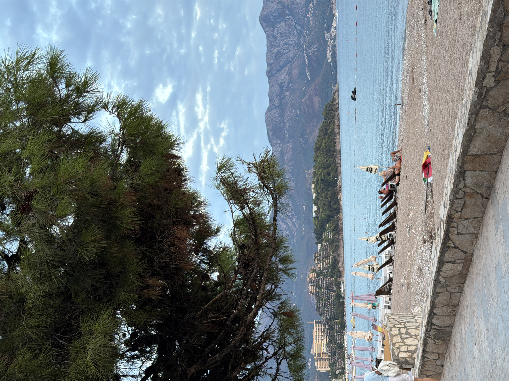 The image depicts a scenic view of a beach in Budva, Montenegro, likely near the Budva Riviera, with the Adriatic Sea in the foreground and mountains in the background. The perspective is from a paved walkway with a stone wall bordering a pebbly beach. Overhanging pine branches with visible cones frame the upper portion of the image.
On the beach, several lounge chairs are set up, some occupied by individuals relaxing. Beach umbrellas, some open and some closed, are scattered across the sand. A few people are visible on the beach, with one person lying on a towel. The sea is calm and blue, with a jet ski visible in the distance, marked by buoys.
Across the water, a coastline is dotted with buildings, including hotels and residential structures nestled into the hillsides. The architecture suggests a resort town. The background features a mountain range with rugged terrain, partially covered in vegetation. The sky is overcast with diffused clouds, indicating either early morning or late afternoon light. The overall impression is a peaceful coastal setting.