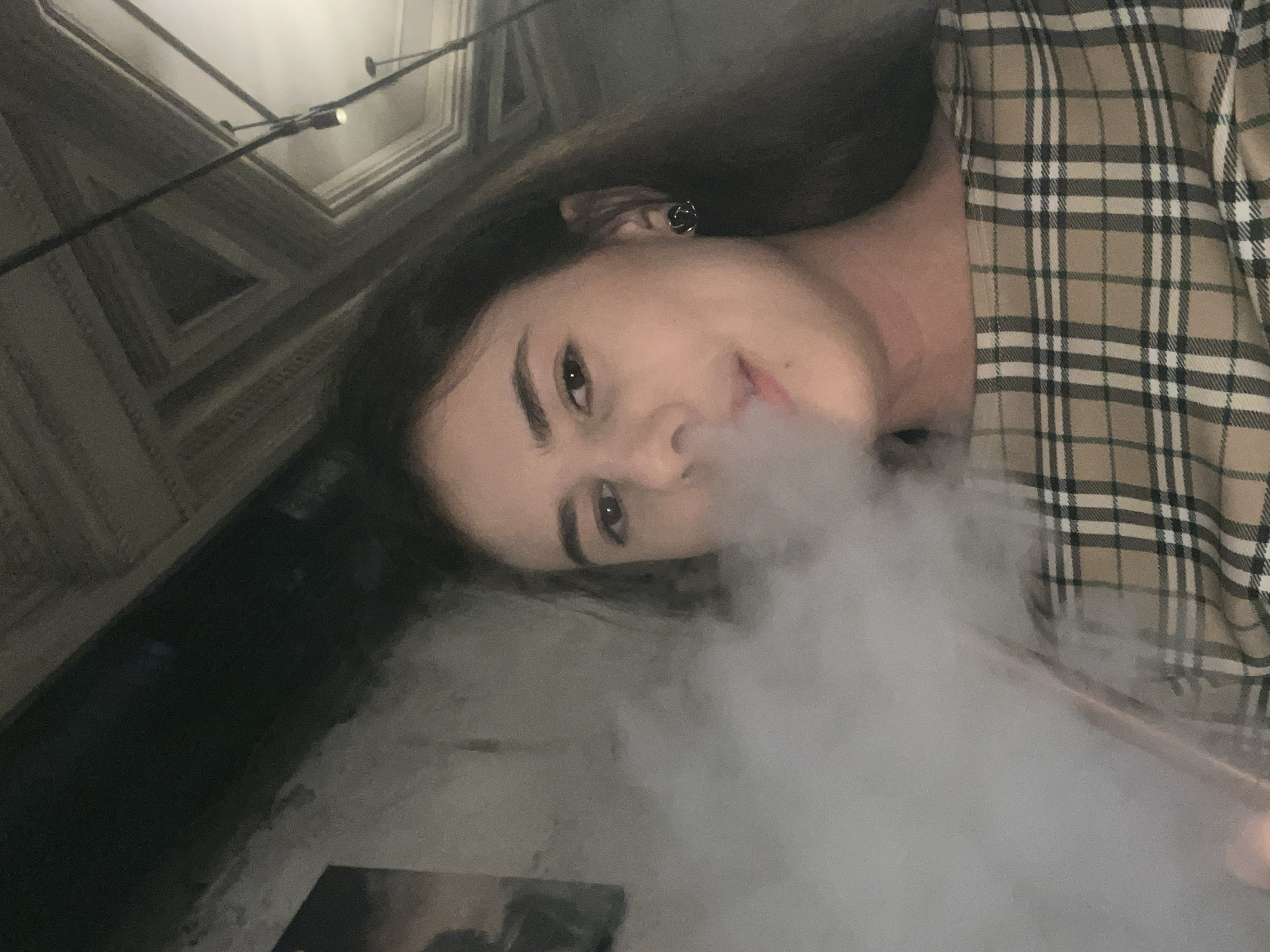 In a dimly lit indoor setting in Unknown, Unknown, a young female is positioned in the foreground, exhaling a dense cloud of white vapor from her mouth. She has long, dark hair and is wearing a beige plaid top with black and white accents. A dark, possibly metallic, earring is visible in her right ear. The background reveals an ornate ceiling with complex geometric patterns in muted tones, from which dark pipes or structural elements extend, along with a single illuminated pendant light fixture on the upper right. To the left, a light-colored, possibly textured wall features a partially visible dark rectangular object.