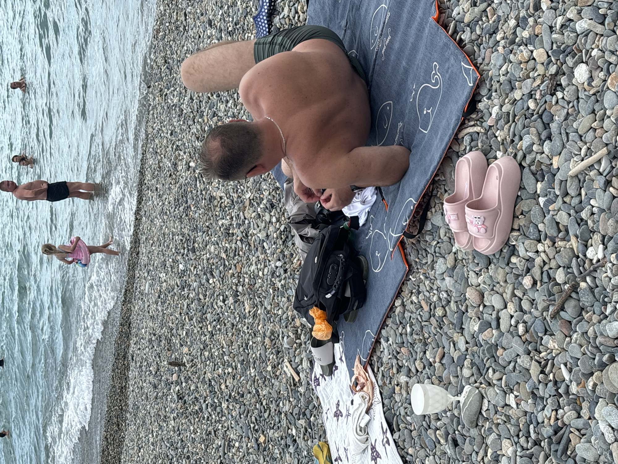 The image depicts a sunny day at a pebble beach in Sochi, Russia. The scene is dominated by a man lying on a towel on the pebble shore. He is wearing dark green swim trunks and a silver chain. His back is to the camera, and he appears to be relaxed. Nearby, a pair of pink flip-flops with a teddy bear design are placed on the pebbles.
In the background, the sea is visible with gentle waves. Several people are in the water, including a man in black swim trunks standing near the shore and a young girl in a pink dress. The water is a muted blue-green.
On the towel with the man, a black backpack and some clothing are scattered. A small orange plastic bag is also visible. Part of another towel with a pattern of stars and fish lies on the pebbles next to the backpack. A clear plastic glass is placed on the pebbles, suggesting a beverage was or is being consumed.
The overall atmosphere is one of leisure and vacation. The lighting suggests it is daytime, likely afternoon, given the strong sunlight. The pebbles are typical of the Black Sea coast.
