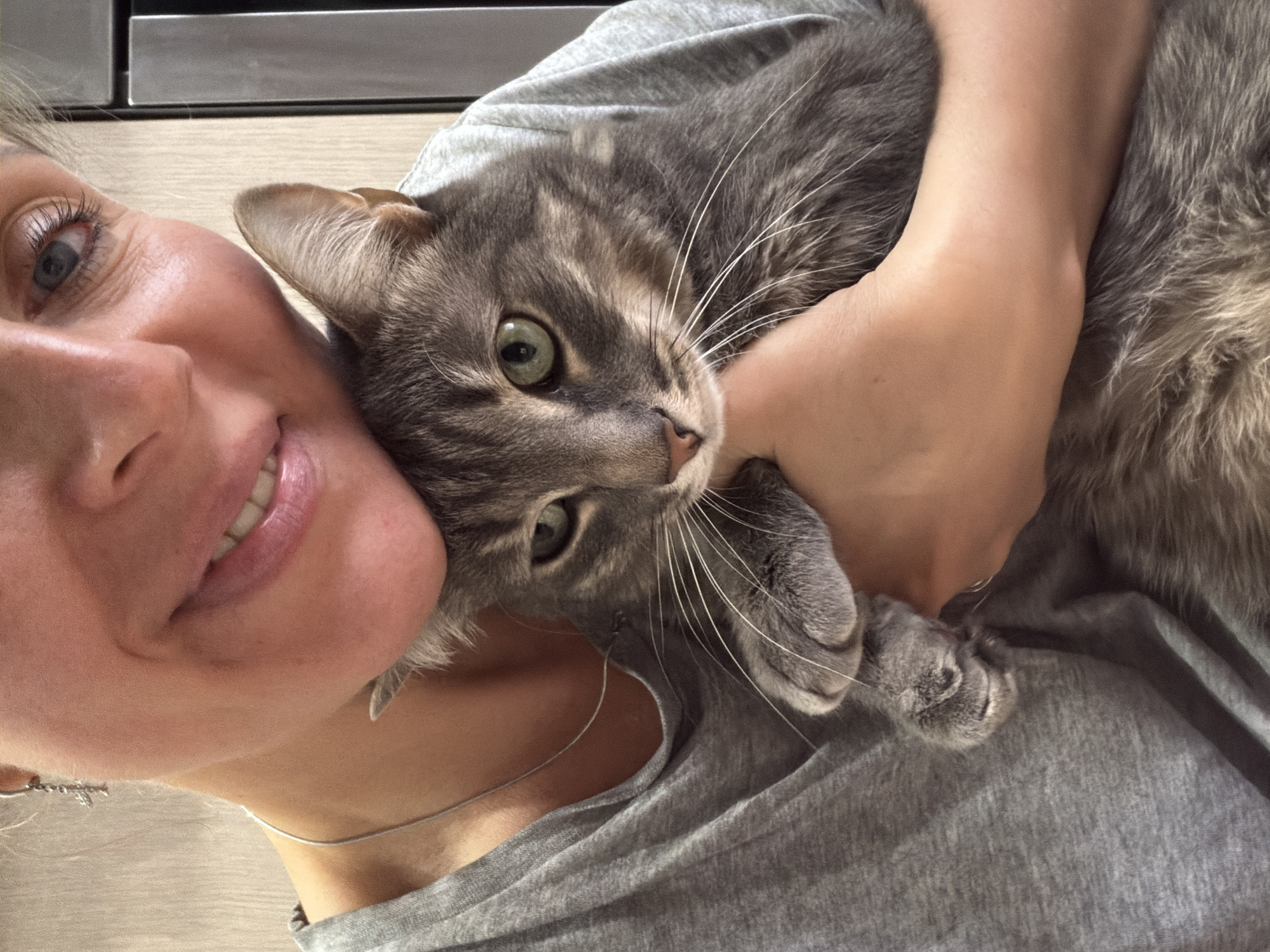 A selfie shows a smiling woman with light skin and blue eyes holding a gray tabby cat with green eyes. The woman is wearing a gray t-shirt and a silver necklace. The cat is nestled in her arms. The background is blurry and appears to be an indoor setting with a light-colored surface. The location in the city of Chaikovsky, Russia, cannot be verified from the image.