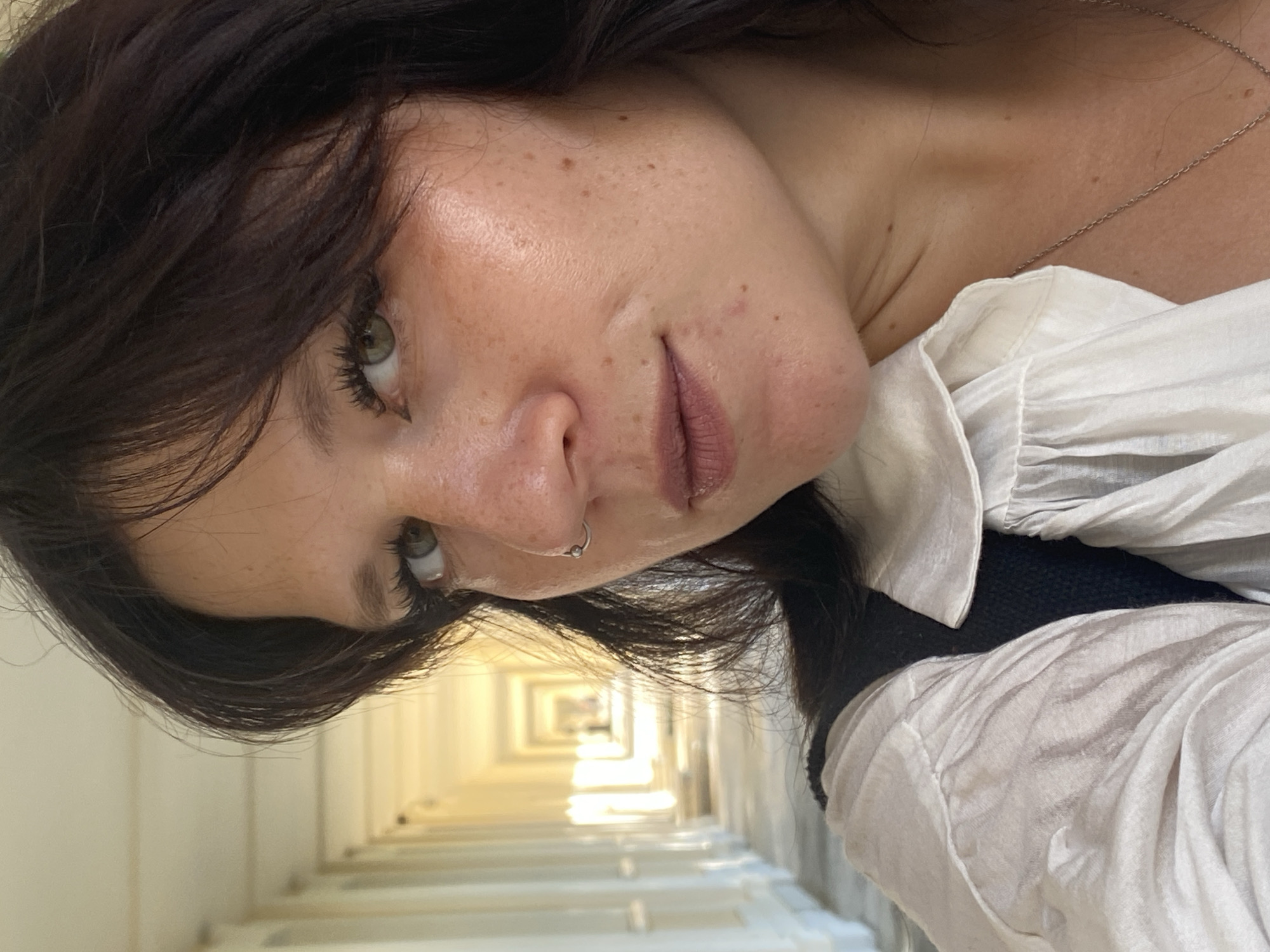 The image is a close-up, profile shot of a woman with dark brown hair, green eyes, and fair skin with freckles. She is wearing a white collared shirt and a dark vest. She has a nose ring. The background appears to be a long, colonnaded walkway, possibly an architectural feature in a park or public building. The lighting suggests it is daytime, with sunlight casting long shadows. The woman is looking towards the camera. The scene is set in the city of Lomonosov, Russia.