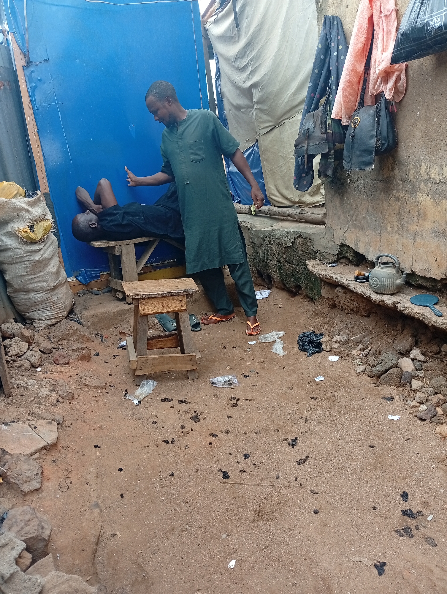 This image captures an outdoor scene in Adankolo, Nigeria, featuring two adult men interacting in a rustic environment. The ground is sandy earth, scattered with rocks, small pieces of plastic trash, and dark, dried debris.
On the left, a man dressed in a dark green tunic and trousers, wearing orange flip-flops, stands over another man. The standing man holds a small, round green and gold object in his left hand, while his right hand is extended towards the chest or shoulder area of the man lying on a simple wooden bench. The man on the bench is dressed in a dark shirt and shorts, resting on his back with his head to the left. Behind the bench is a bright blue wall or tarp. To the far left, a large, light-colored sack filled with an unknown substance leans against this blue structure.
To the right of the scene, a rough, earthy-toned wall is visible. Several items are hanging from it, including multiple dark-colored handbags, a polka-dotted fabric, and a light peach-colored fabric. Below, on a concrete ledge, sits a grey, ridged teapot or kettle, a small circular container, and a blue hand mirror. A small wooden stool is placed in front of the bench, and a single black slipper lies on the ground beneath it.
The natural lighting suggests it is daytime. The informal structures, dirt ground, and everyday objects point to a humble, perhaps informal commercial or residential setting. The only visible text, "VIVA," is faintly printed on a black plastic bag hanging on the wall. The interaction between the two men appears focused and calm.