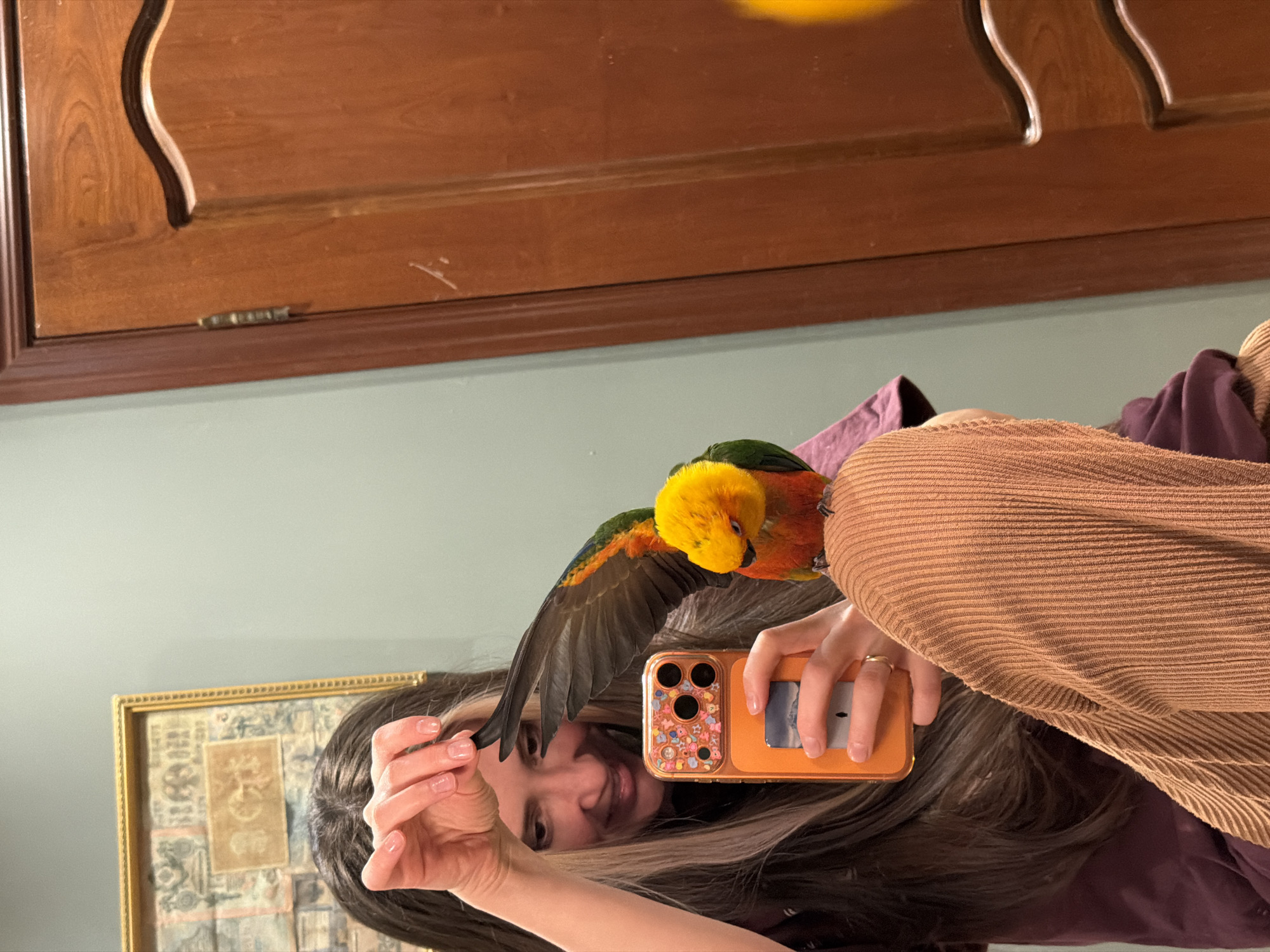 A woman is holding an orange smartphone with a decorative case, seemingly taking a selfie. A Sun Conure parrot is perched on her shoulder. The woman has long brown hair and is wearing a brown corduroy shirt. The background includes a framed picture and a wooden cabinet. The image does not depict the city of Khimki, Russia.