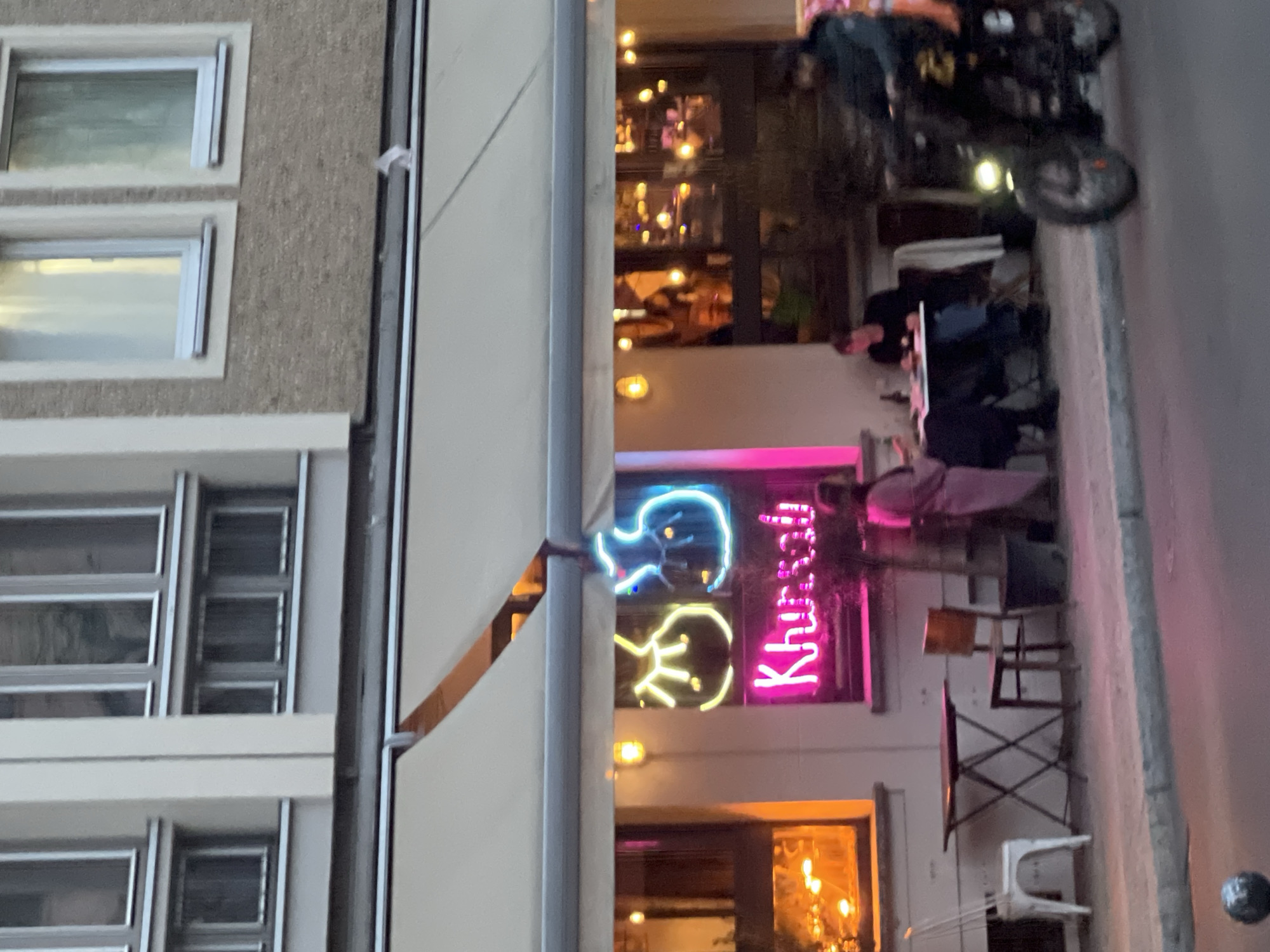 The image depicts an evening street scene in Berlin, Germany, focusing on the exterior of a building, likely a restaurant or bar, with outdoor seating. The perspective is slightly angled, looking towards the establishment from across the street.
Several people are visible. On the right side, two individuals are seated at an outdoor table, appearing to be dining or socializing. Another person is partially visible further inside the seating area. At the top right, a person is on a motorcycle, which is either parked or in motion on the street.
The building itself features multiple rectangular windows with light-colored frames set into a textured, possibly pebbled, facade. A light-colored awning extends outwards, covering part of the sidewalk seating area.
A prominent feature is a vibrant neon sign on the building's facade. In bright pink neon, the text "Khúc Cây" is clearly visible. Next to this text are two stylized neon figures or logos, one in blue and one in yellow, resembling abstract heads or faces. The interior of the establishment glows with warm lights, suggesting activity within. Various outdoor tables and chairs, including a white plastic chair and metal folding chairs, are arranged on the pavement.
The illuminated neon signs and interior lights indicate that the scene takes place during dusk or evening. The atmosphere appears social and relaxed.