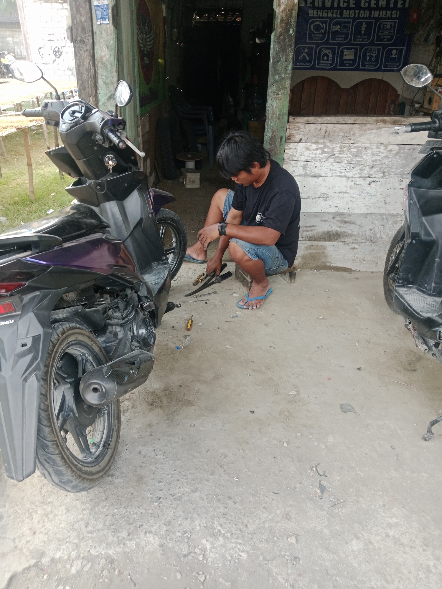 In the city of Trate, Indonesia, a mechanic in a black t-shirt and denim shorts is seen crouched on the ground, working on a purple and black scooter. He is holding a tool, possibly a wrench, and appears to be in the process of repairing the scooter. The scooter is parked next to another black scooter. The scene is set in an outdoor workshop with a wooden building in the background displaying a sign for "SERVICE CENTER BENGKEL MOTOR INJEKSI". Tools and parts are scattered around the mechanic, indicating ongoing work. The ground is a dusty concrete surface, with some gravel and debris.