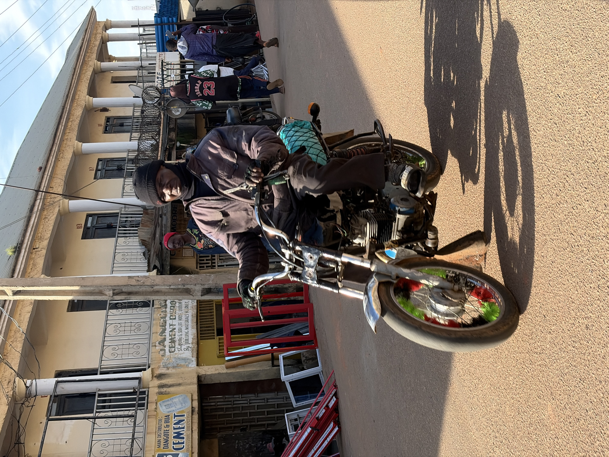 A man wearing a dark beanie and a brown jacket rides a motorcycle on a dusty road in Gura Top, Nigeria. The motorcycle has a blue woven bag strapped to the back. In the background, a two-story building with balconies and pillars is visible, along with several shops displaying red and white window frames and building materials. The sun casts long shadows on the road, indicating it is late afternoon or early morning.