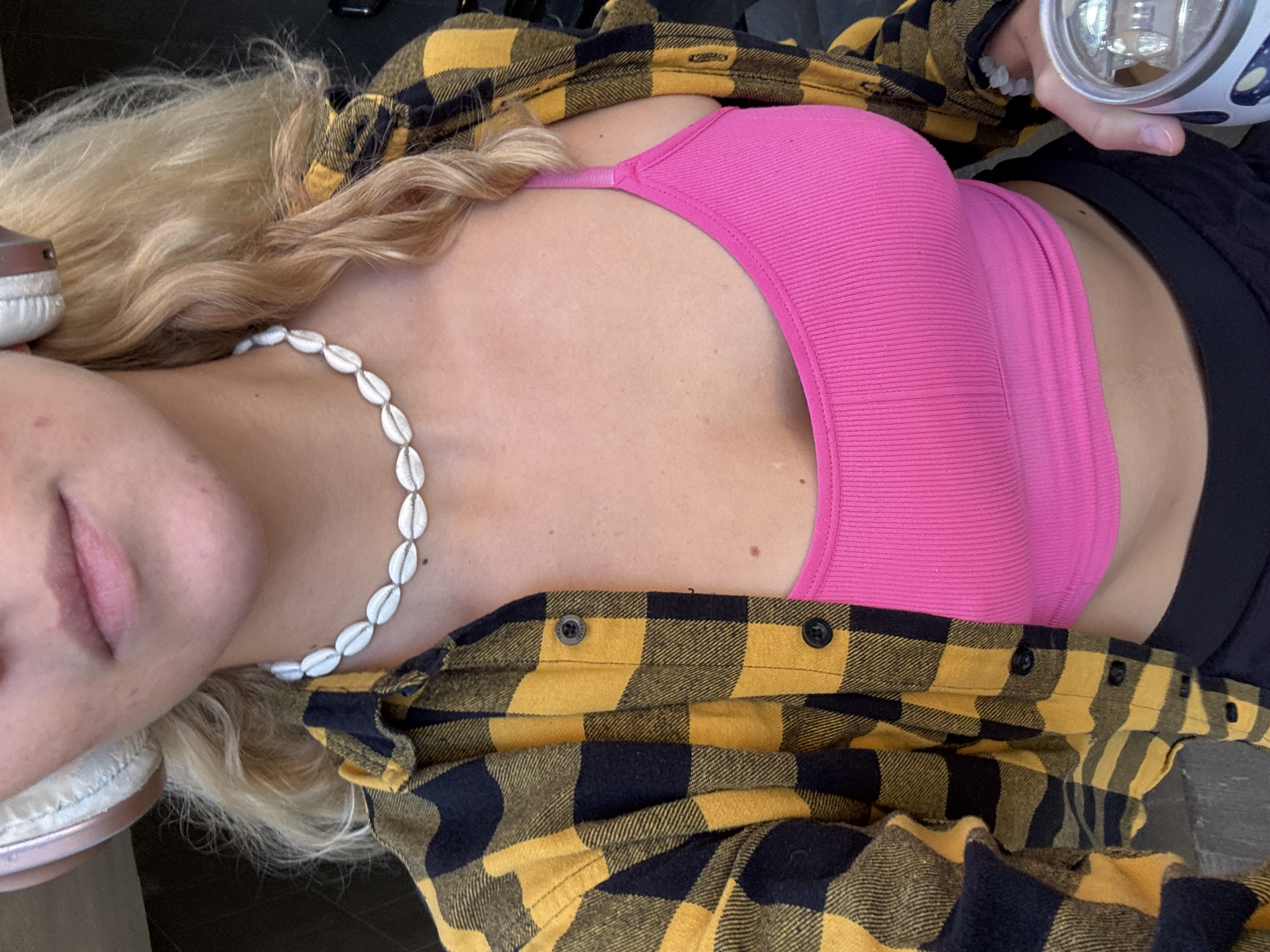 This close-up, eye-level shot features a person, likely female, from the chest up, oriented diagonally with their head positioned towards the upper left. The person has long, wavy blonde hair and is wearing a bright pink ribbed tank top or sports bra layered under an unbuttoned yellow and black buffalo plaid flannel shirt.
A white cowrie shell necklace adorns their neck, resting on their chest. White over-ear headphones are visible near their left ear and shoulder. The left side of their face, including the chin and partially parted lips, is visible. In the upper right corner, a hand holds a clear, cylindrical object with a white and blue base, which could be a water bottle or a portable device. The background is dark and indistinct, suggesting an indoor or shaded setting. The overall scene conveys a casual atmosphere. There is no clearly legible text on any visible items. The context provided indicates the location is Minsk, Belarus.