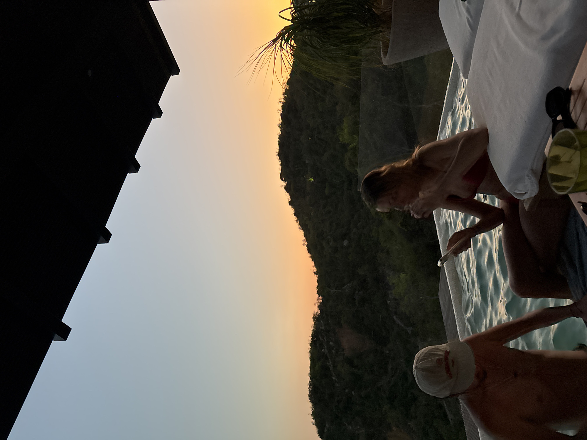 The image shows a sunset scene at a pool, likely in a tropical or coastal location.
People: A woman in a red swimsuit is visible, holding a smartphone. A man wearing a white cap is in the pool.
Objects: Pool with light blue water, white lounge cushion, green plant, clear glass.
Environment: Green mountains are in the background, and the sky has hues of orange and blue, indicating sunset. The location may be in Puerto Marqués, Mexico, given the foliage and the general landscape. The edge of a dark roof is visible on the left side of the image.