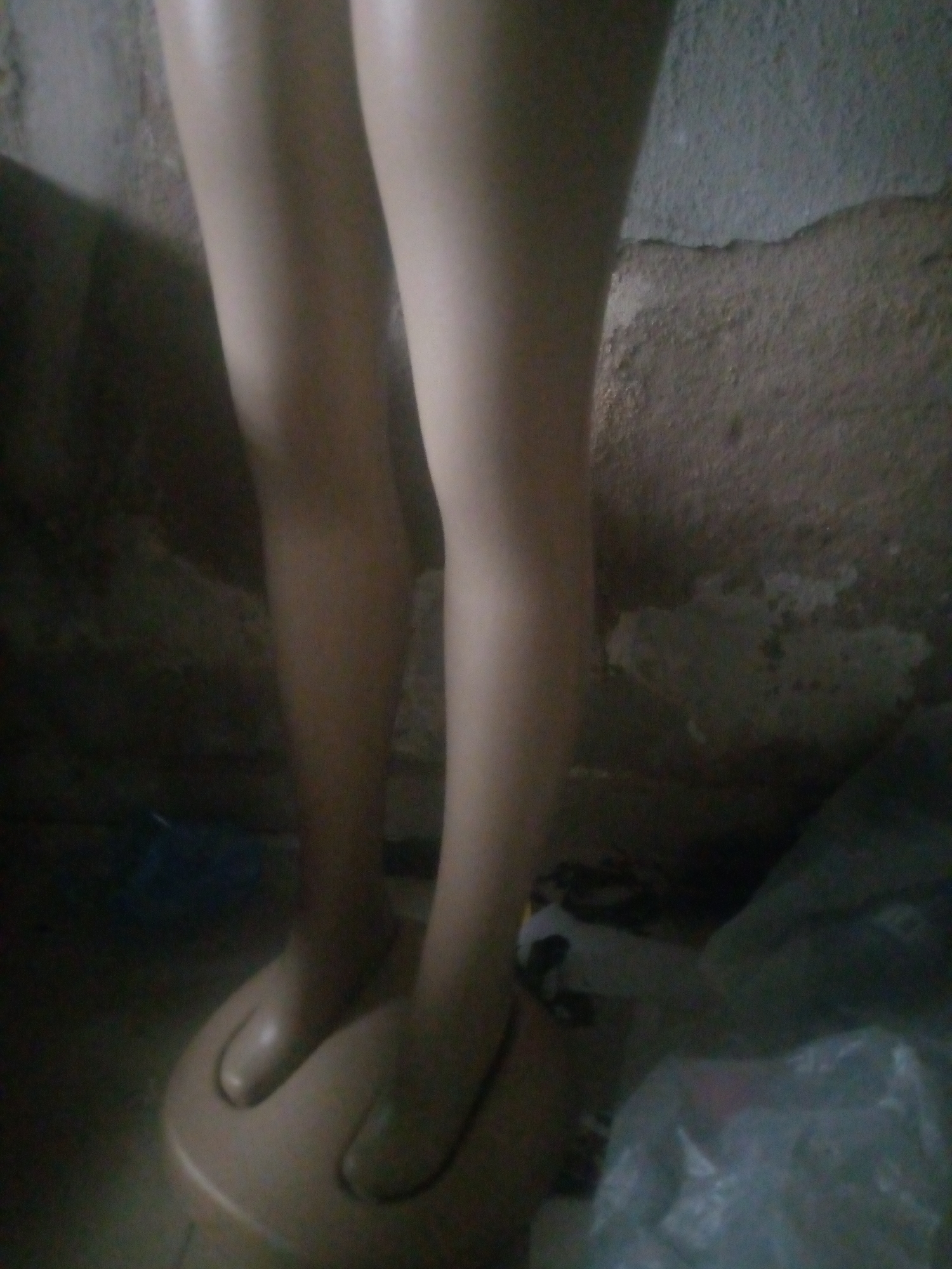 The image shows the lower half of two mannequin legs standing on a round base. The legs are smooth and flesh-toned, with subtle shading. The background is a rough, textured wall, likely made of concrete or stucco, with a lighter colored patch towards the right side. The floor is dark and appears to be cluttered with some objects, including a clear plastic bag on the right. The lighting is dim, casting shadows and creating a somewhat grainy appearance. The location is identified as Jauro Yinu, Nigeria.