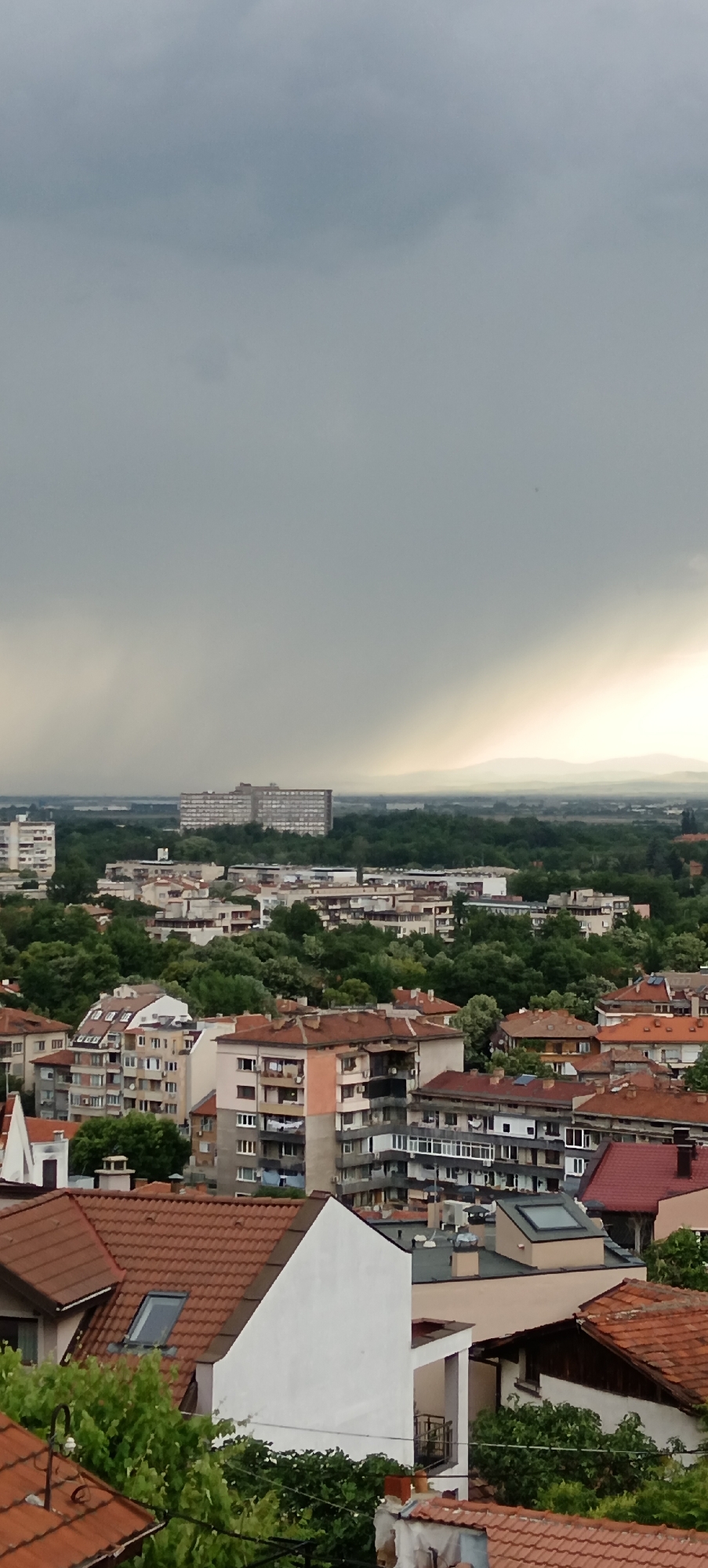 An elevated view captures a city landscape under a dramatic sky, possibly Plovdiv, Bulgaria. The foreground features residential rooftops with terracotta tiles, intermixed with foliage. Mid-ground reveals apartment buildings surrounded by greenery, transitioning into a skyline with a large, rectangular building in the distance. The sky dominates the upper half of the image, with a grey, stormy appearance and a patch of lighter area near the horizon. Distant hills are barely discernible on the horizon.