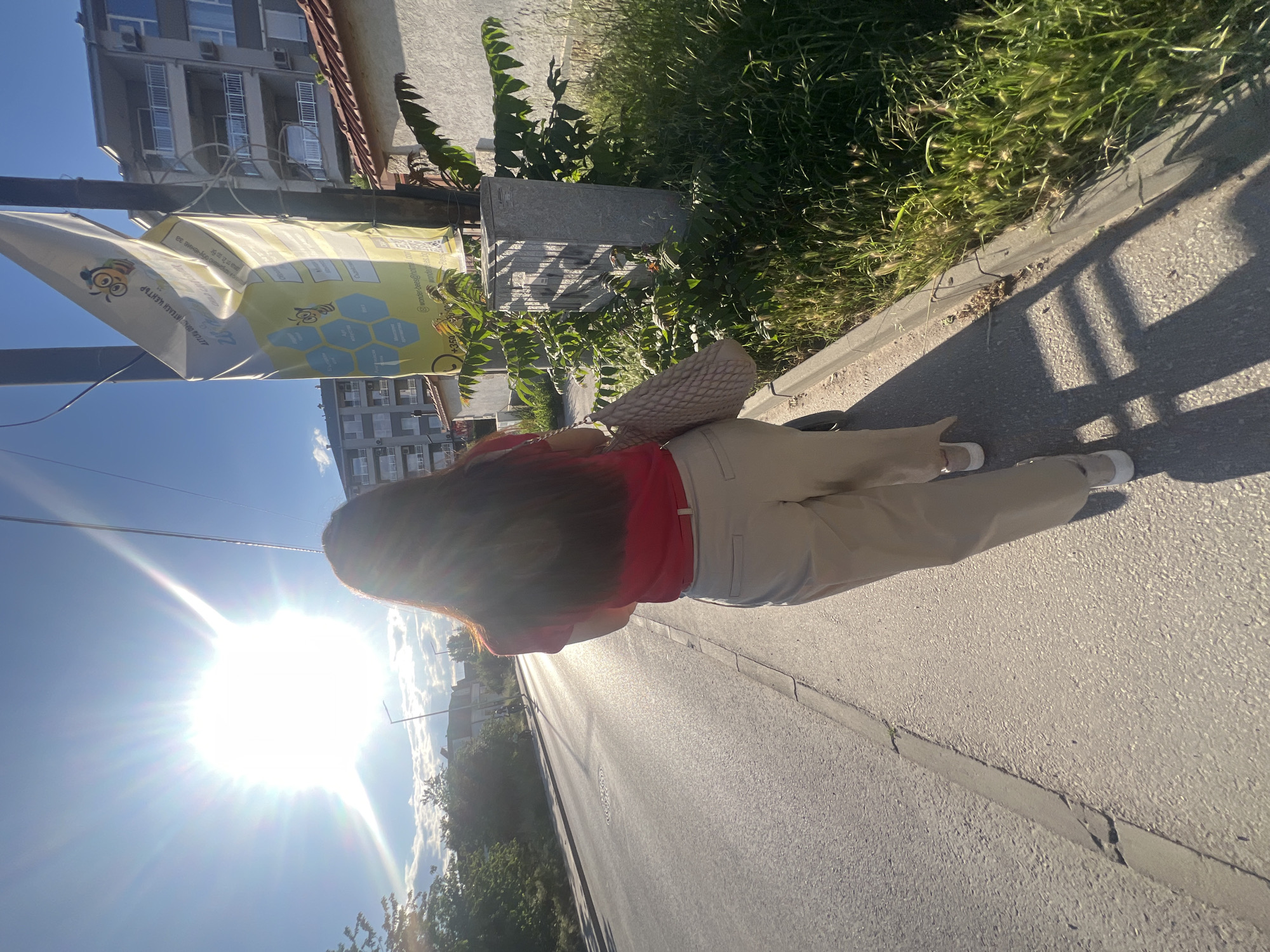 The image, likely taken in the city of Plovdiv, Bulgaria, shows a view of a person from behind, facing away from the camera, walking along a city street on a sunny day.
**People:** The person is wearing a red top and beige pants, with their brown hair flowing down their back. They are carrying a beige handbag.
**Objects:** The image captures a street scene with a building, sidewalk, street vegetation, and a street lamp post, and a sign attached to the post is visible.
**Environment:** The scene is set in an urban environment with buildings, roads, sidewalks, and vegetation. It is a sunny day, as indicated by the bright sunlight and shadows. The camera position is from a high angle, looking down on the individual walking along the street.