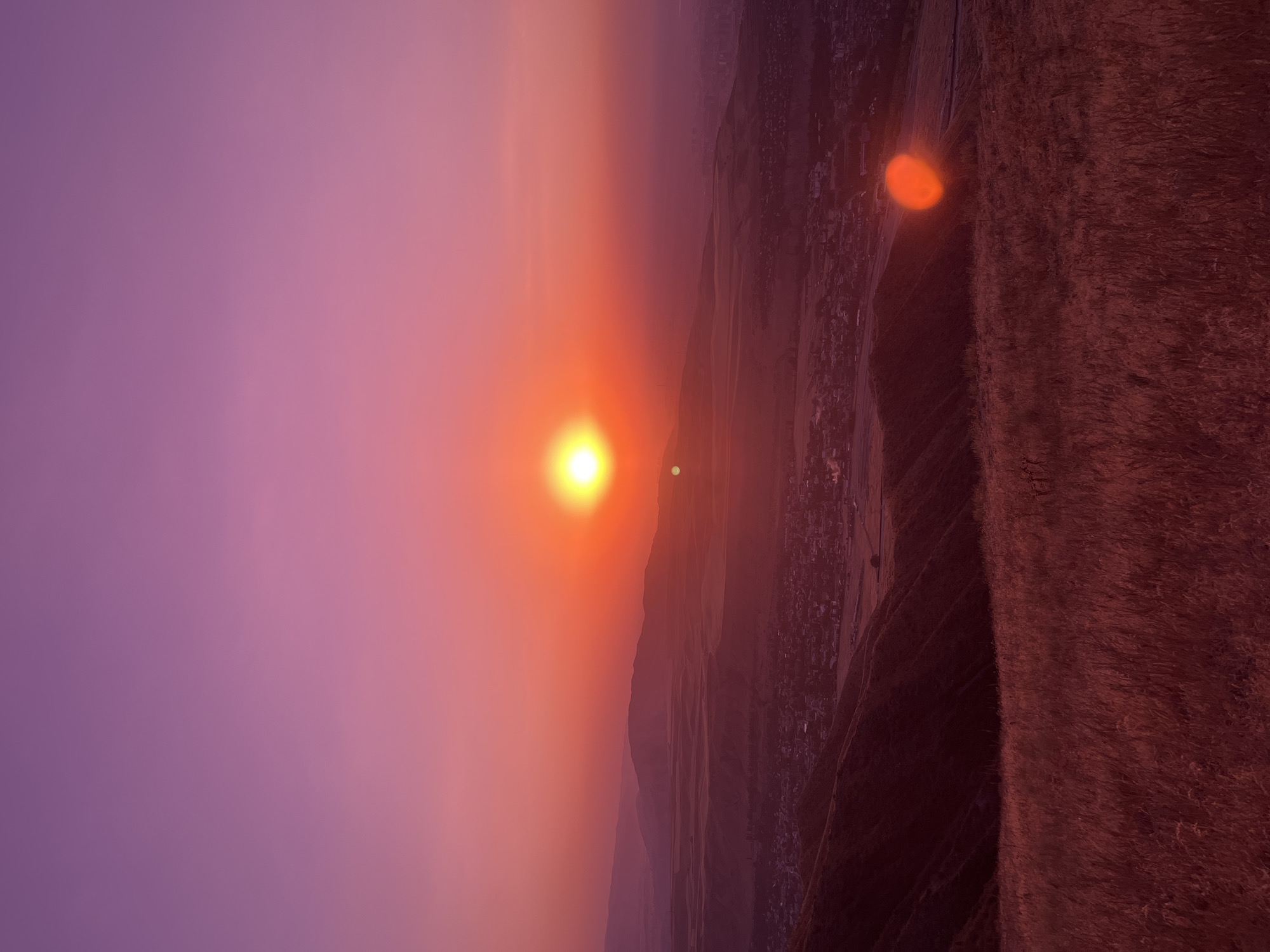 A high-angle, eye-level, medium shot captures a panoramic view of a sunset over Besh Kungei, Kyrgyzstan. The sky transitions from a deep purple at the top to a vibrant orange and red as it nears the horizon, painted by the setting sun. The sun, a bright, glowing orb, is positioned in the upper-middle portion of the frame, casting a warm light across the landscape.

Below the sky, rolling hills and mountains form the middle ground, their contours softened by the fading light. A town is visible nestled in the valley, its buildings appearing as small, dark shapes. The foreground is dominated by a field of dry, golden grass, its texture enhanced by the sunset's glow. A prominent lens flare, a bright orange oval, appears in the lower right corner of the image, a byproduct of the sunlight interacting with the camera lens. The overall scene evokes a serene and tranquil atmosphere, a testament to the natural beauty of the Kyrgyz landscape at dusk.