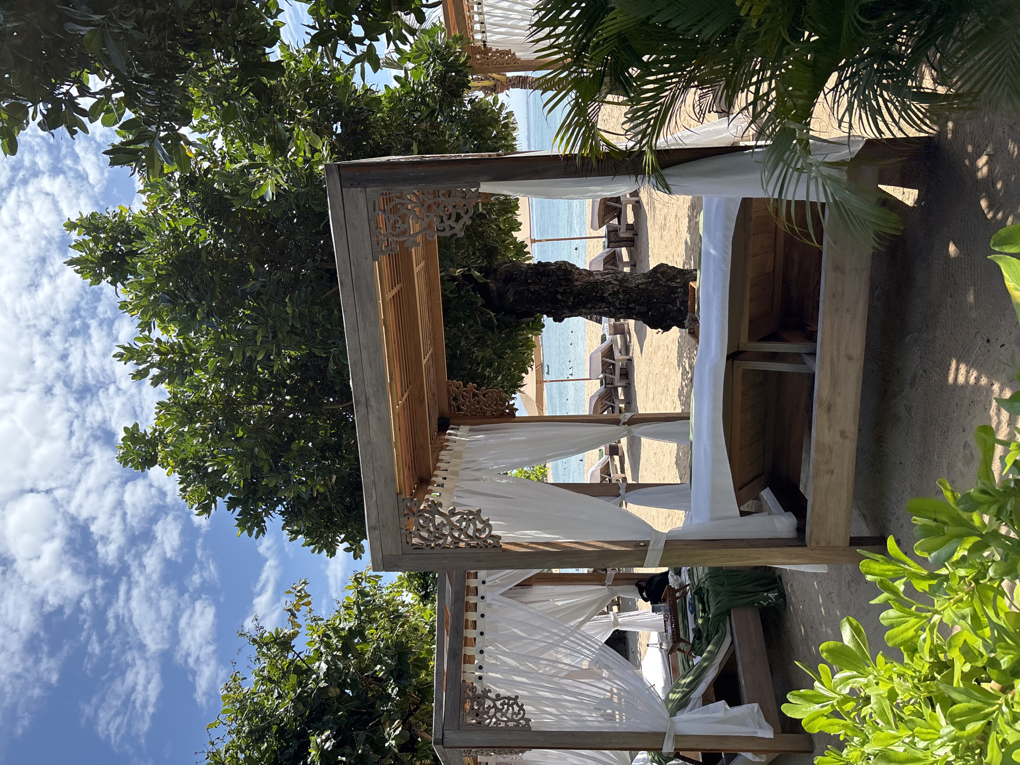 The image presents a scene on a beach in Kuta Selatan, Indonesia, featuring a wooden cabana with white curtains. Through the cabana, the ocean and other beach loungers can be seen. A tree is visible behind the cabana, and foliage surrounds it.