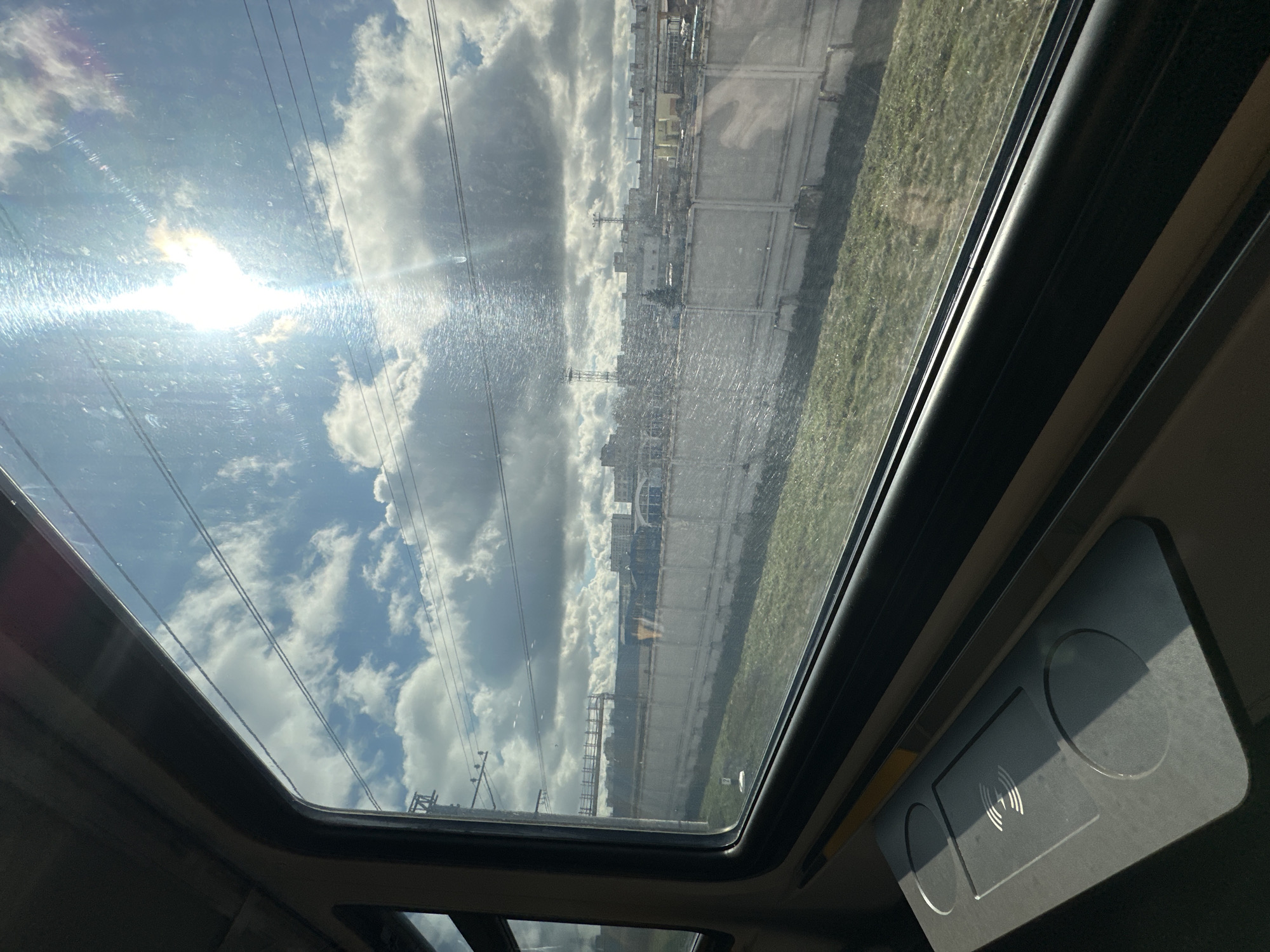 The image is taken from the interior of a vehicle, looking up through a sunroof. Bright sunlight and clouds are visible through the glass, with power lines cutting across the sky. In the background, beyond the clouds, is an urban industrial area with tall buildings and a concrete wall. The right side of the image shows part of the car's interior, including what appears to be a wireless charging pad. Given the architecture and infrastructure style visible through the sunroof, the location is likely in or near Moscow, Russia.