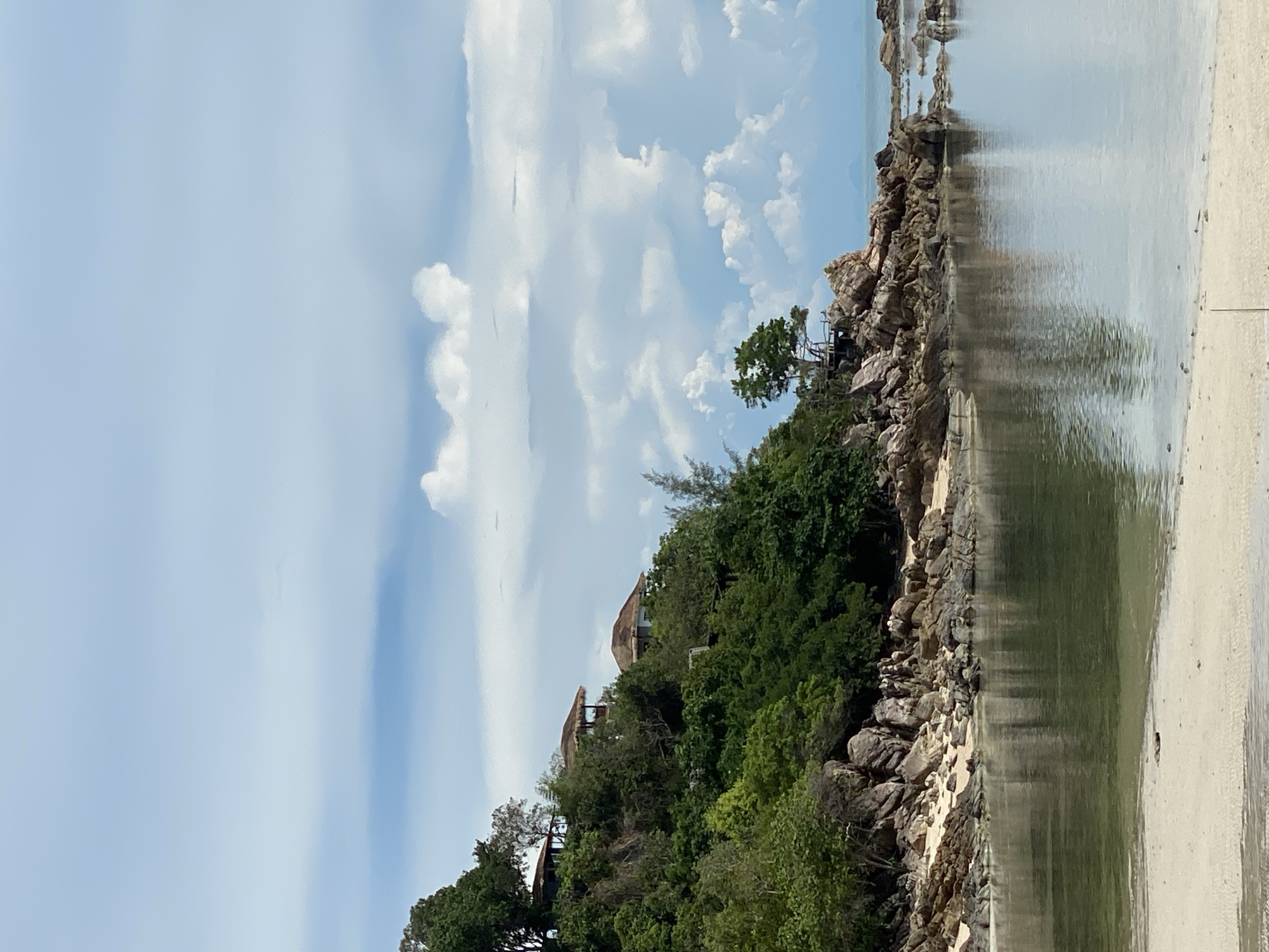 This is a vertical, eye-level shot of a tropical coastline on Koh Phangan, Thailand, under a cloudy sky. The foreground features a sandy beach leading into calm, reflective water. The water is a muted green and reflects the sky and surrounding foliage. The midground is dominated by a rocky shoreline with sparse vegetation and scattered boulders. Beyond the rocks, a densely vegetated hillside rises, covered in lush green trees and shrubs. Two buildings with reddish-brown roofs are visible nestled amongst the trees on the upper part of the hillside, suggesting a resort or residential area. The sky is a pale blue with scattered white and grey cumulus clouds, some of which are large and billowy. The overall atmosphere is serene and natural, with a hint of human habitation integrated into the landscape.
