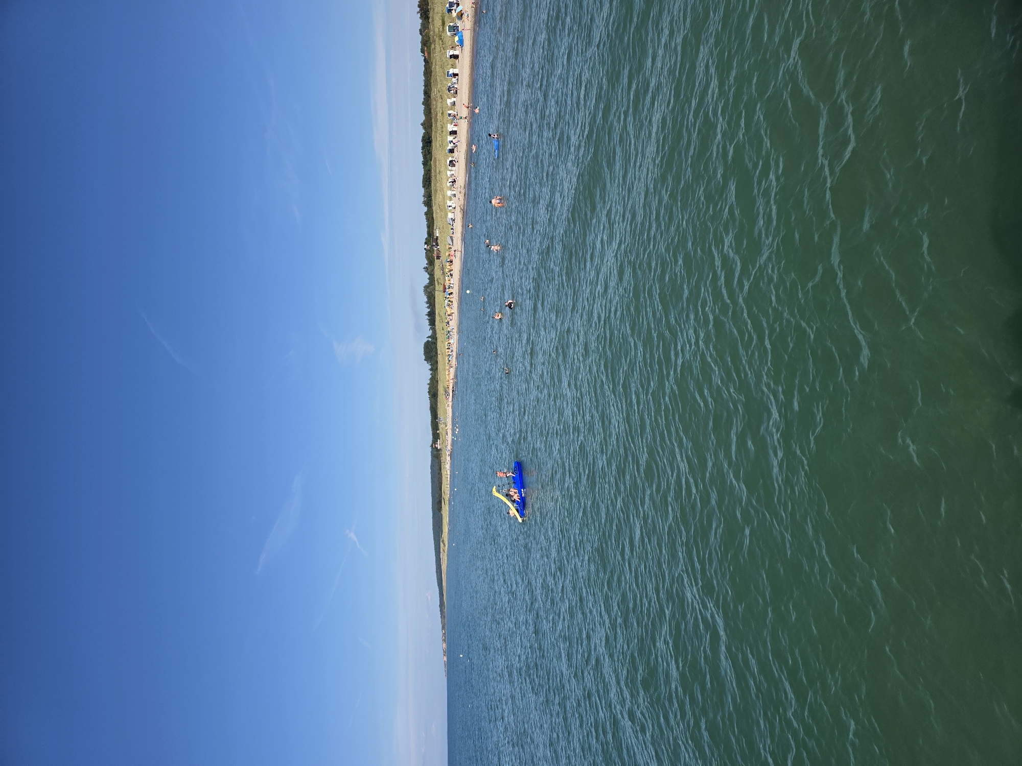 This image shows a sunny day at the beach in Wangels, Germany, specifically at the Brodhaus Bay. The scene is dominated by a vast expanse of calm, green-blue water in the foreground, with gentle ripples on its surface. In the middle ground, a crowded beach stretches along the coastline, lined with numerous white and blue striped beach chairs and umbrellas. People are scattered along the sandy shore, sunbathing and relaxing. Further out in the water, several individuals are swimming and enjoying themselves. A prominent inflatable water toy, a blue structure with a yellow slide, is in the water with a few people on it, some of whom appear to be children. The background features a dense line of trees and vegetation, with a clear blue sky overhead, punctuated by a few wispy clouds.