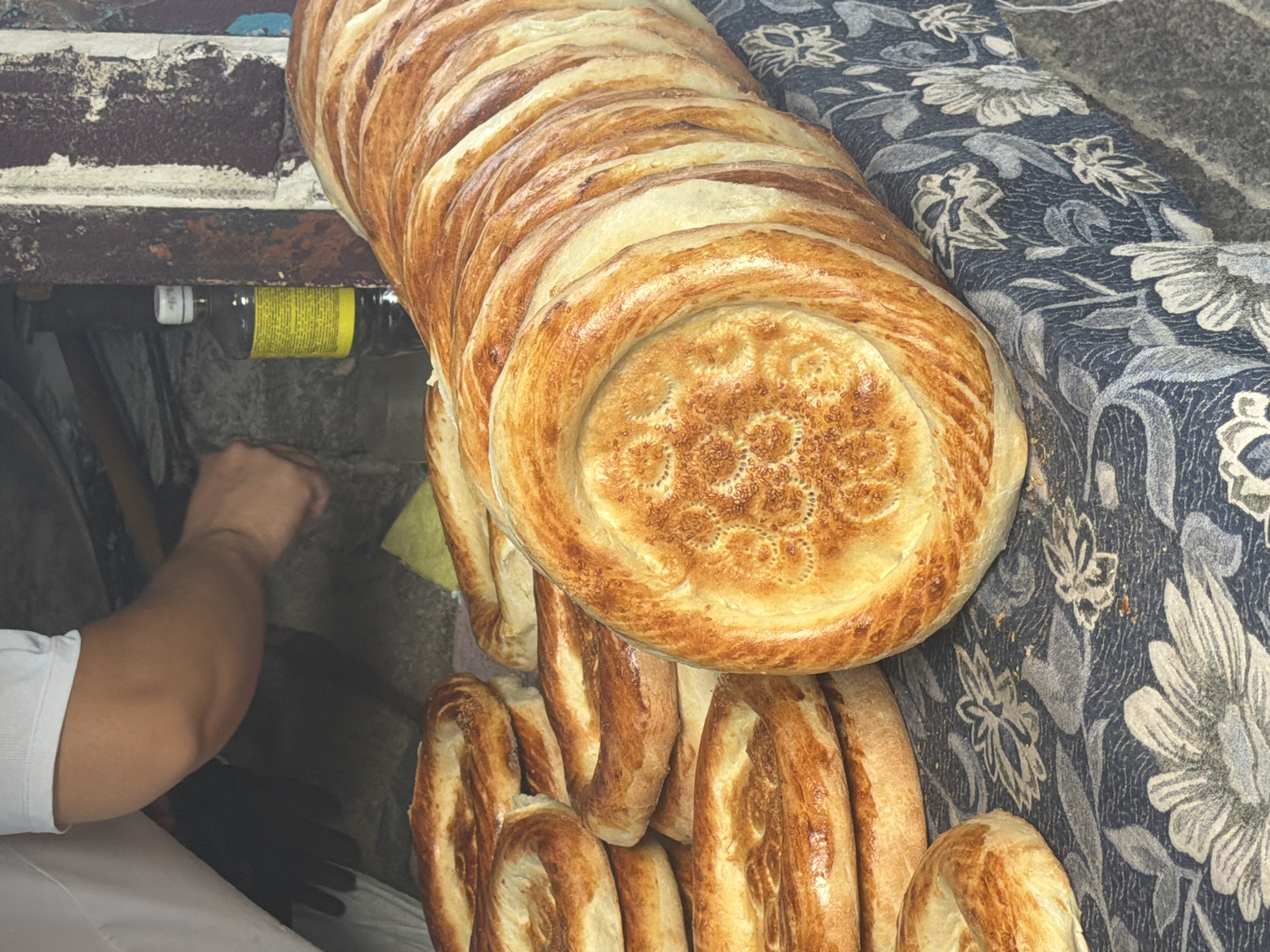 An over-the-shoulder, medium shot captures a stack of freshly baked, circular flatbreads resting on a dark, floral-patterned tablecloth. The bread, golden brown with slightly darker edges, features decorative indentations on its surface. In the background, a person's arm, wearing a white t-shirt, is visible, engaged in an unseen task, possibly preparing more bread. To the right, a dark brown bottle with a yellow label stands upright. The setting appears to be an outdoor market stall or bakery.