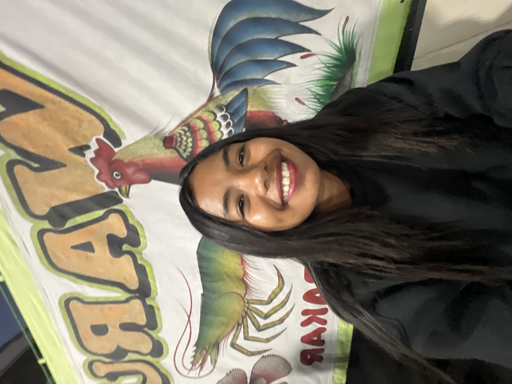 In a nighttime outdoor scene, a young woman with long, dark hair smiles broadly at the camera. She is wearing a black sweatshirt. Behind her, a banner depicts a large green shrimp and a colorful rooster, with large, bold lettering in yellow and orange with black outlines above them. Below the shrimp and rooster, red lettering is visible on the banner. The context suggests this is a food stall or restaurant in Denpasar, Indonesia, likely at night given the lighting and the presence of the illuminated banner.