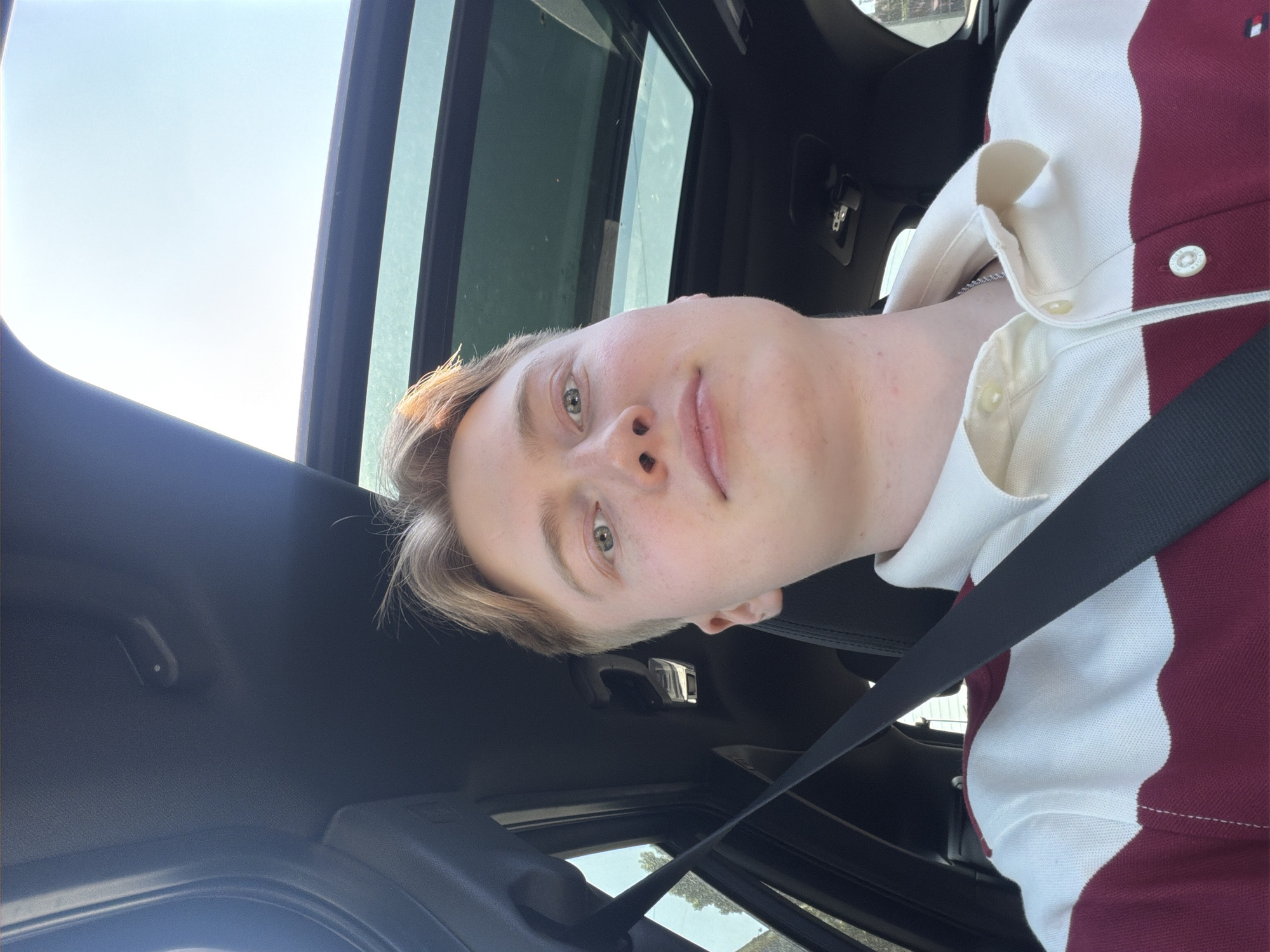A young male with light brown hair is seated in the passenger seat of a car, looking directly at the camera. He is wearing a polo shirt with horizontal stripes of white and burgundy. A black seatbelt is visible across his chest. The car has a sunroof, which is open, allowing sunlight to illuminate his hair. The interior of the car, including the ceiling and a portion of the dashboard, is visible. The background shows a bright sky and faint greenery, suggesting an outdoor environment. There is no discernible information within the image that specifies a location in Kyiv, Ukraine.