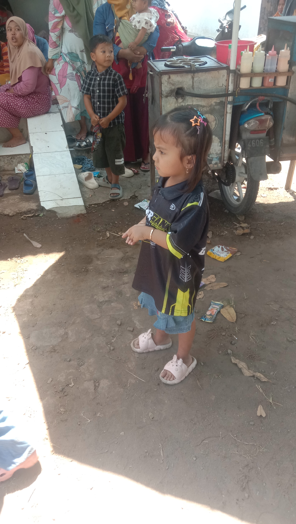 A medium shot captures a young girl in a black and yellow shirt and denim shorts, standing on a dirt ground in Ngempit, Indonesia. She is facing right, looking off-camera, with star-shaped hair clips in her dark hair and a gold bracelet on her right wrist. In the background, a food cart is visible, with a motorcycle attached. Several people are present, including a woman in a pink hijab, a boy in a plaid shirt, and a person holding a baby. The environment appears to be outdoors, with the ground covered in dirt and scattered leaves. The overall scene suggests a casual, outdoor gathering or market setting.