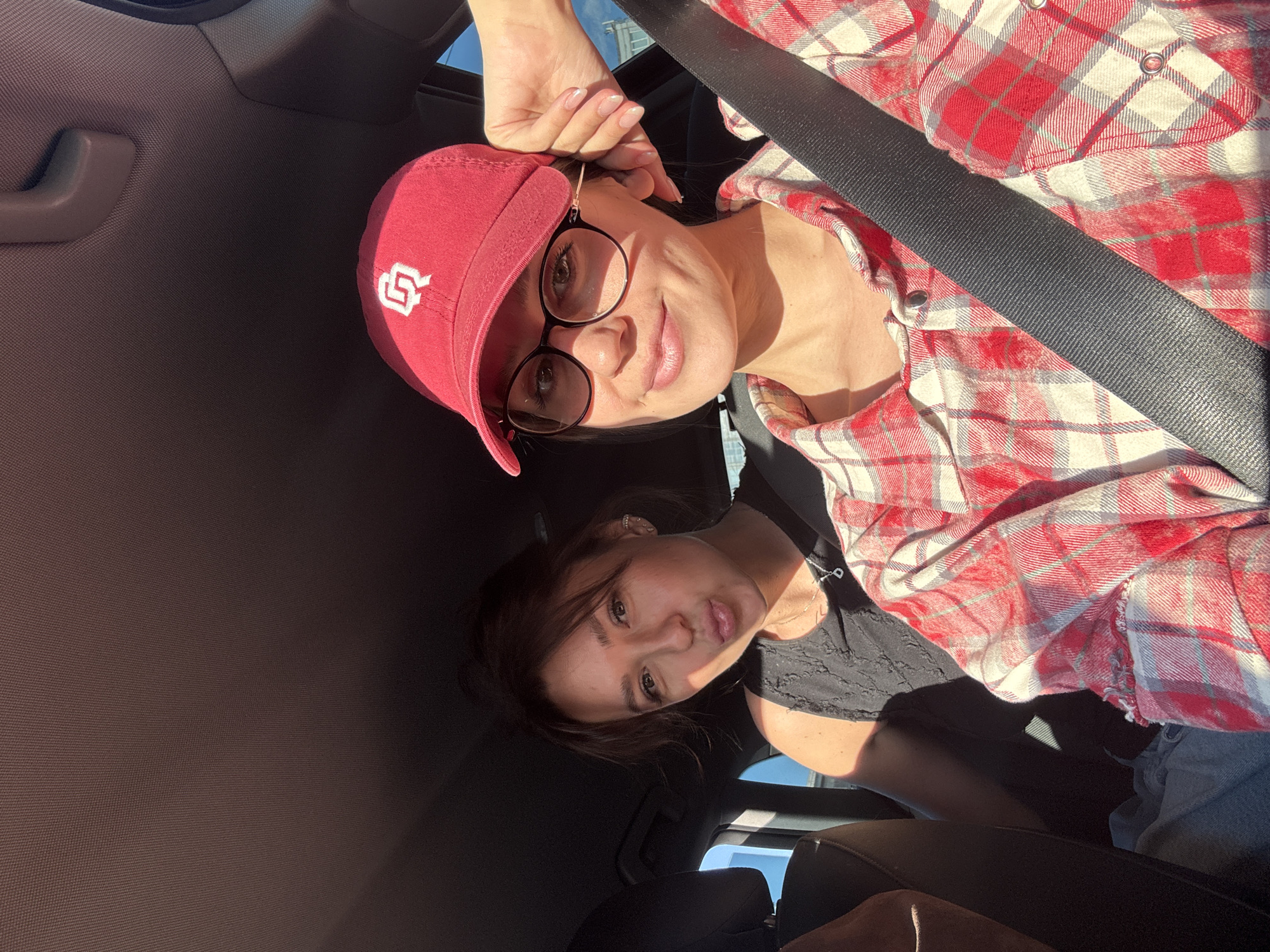 Two women are in a car. One woman is wearing a red and white flannel shirt and a red baseball cap with a white logo. She is also wearing glasses. The other woman is wearing a black shirt. Both women are wearing seatbelts. The car is likely moving as the photo is angled. The location is unknown, and there is no information to suggest the city of Krasnoyarsk, Russia.