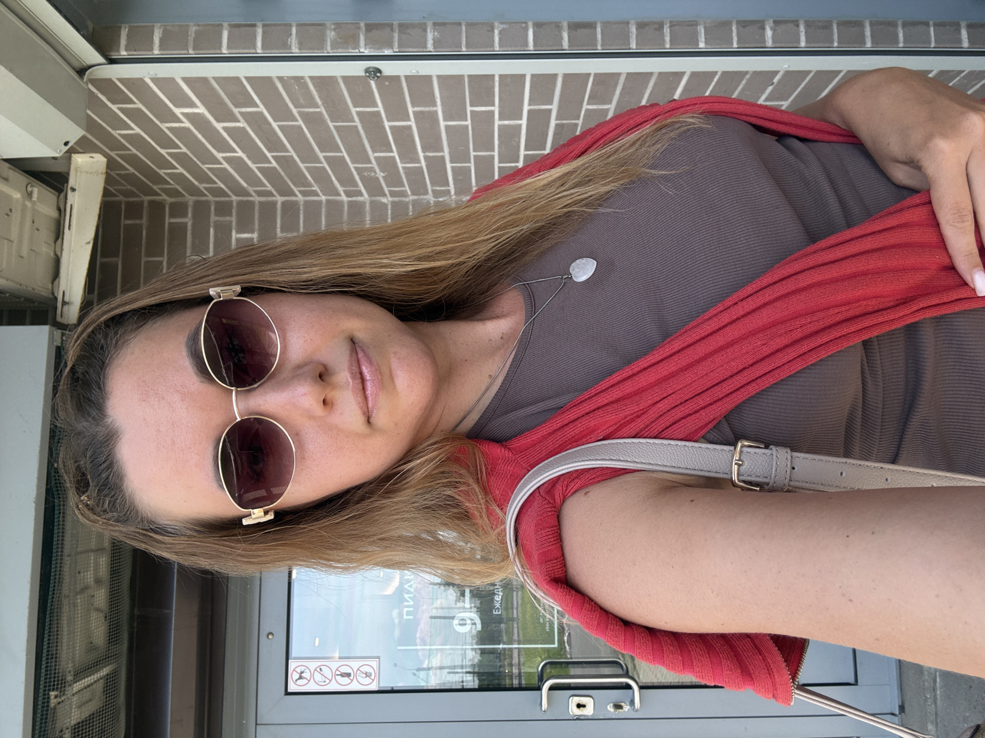 A woman stands in front of a brick building with a glass door. She is wearing sunglasses, a brown tank top, and a red sweater draped over her shoulders. A gray purse hangs on her left shoulder. The door has a sign with several icons on it, and the words "ПИДИ" and "9 ЕЖЕДН" are visible through the glass. The woman is looking at the camera and smiling slightly. The overall scene suggests an outdoor urban environment.