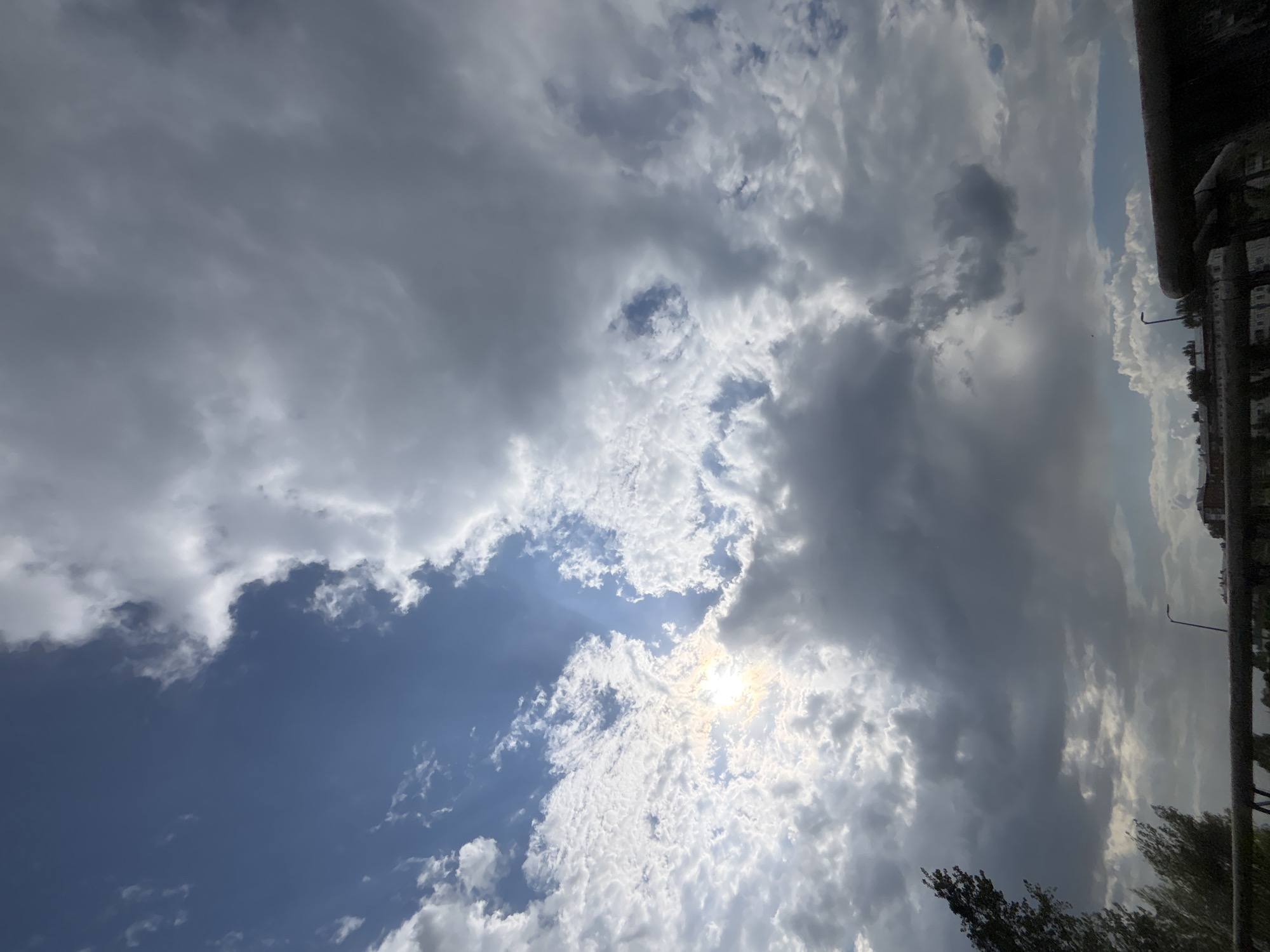 The image shows a cloudy sky above Votkinsk, Russia. There is a visible sun shining through the clouds in the center of the image. The sky is a mix of grey and white clouds with patches of blue. In the lower right corner, parts of a tree and the edge of a building can be seen. A street light is also visible along the building's edge.
