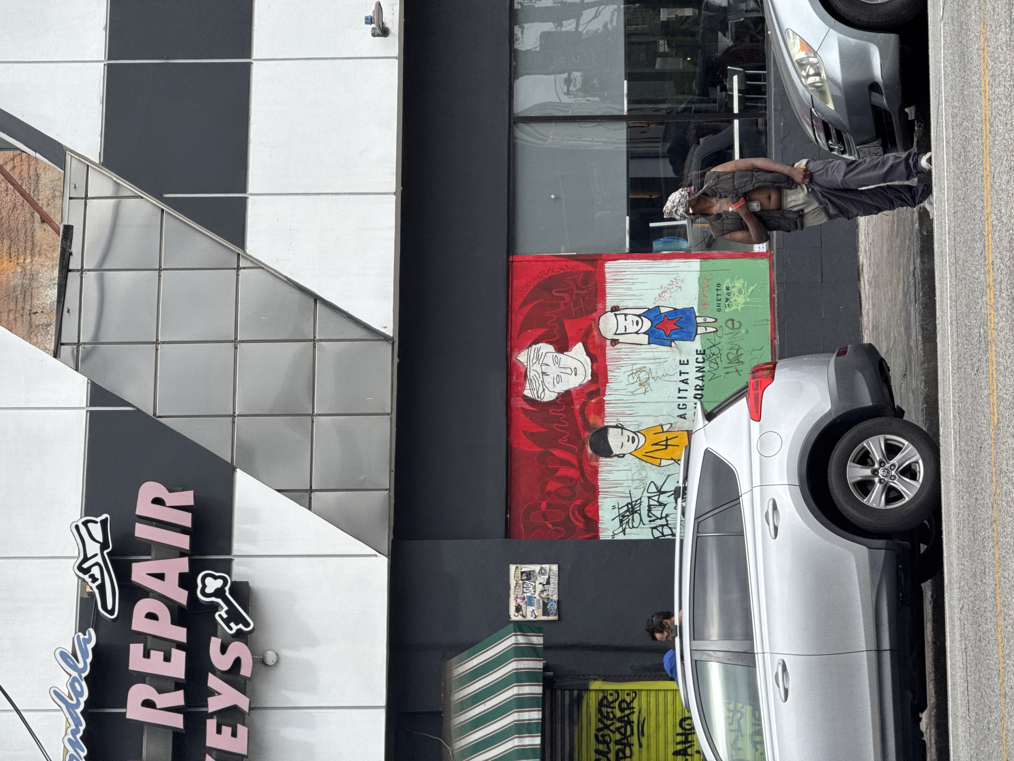 The image shows an urban street scene in Los Angeles, United States of America. On the left, the side of a building is visible with a sign reading "Endola REPAIR KEYS". The building features a geometric pattern of black, white, and silver tiles.
In the center of the frame, there's graffiti art on a wall, depicting stylized faces and writing. A man is standing on the sidewalk next to it.
Two silver vehicles, a car and an SUV, are parked along the curb on the right side of the image, partially obscuring the graffiti. Additional elements in the background include an awning with green and white stripes, and a yellow painted structure with graffiti.
