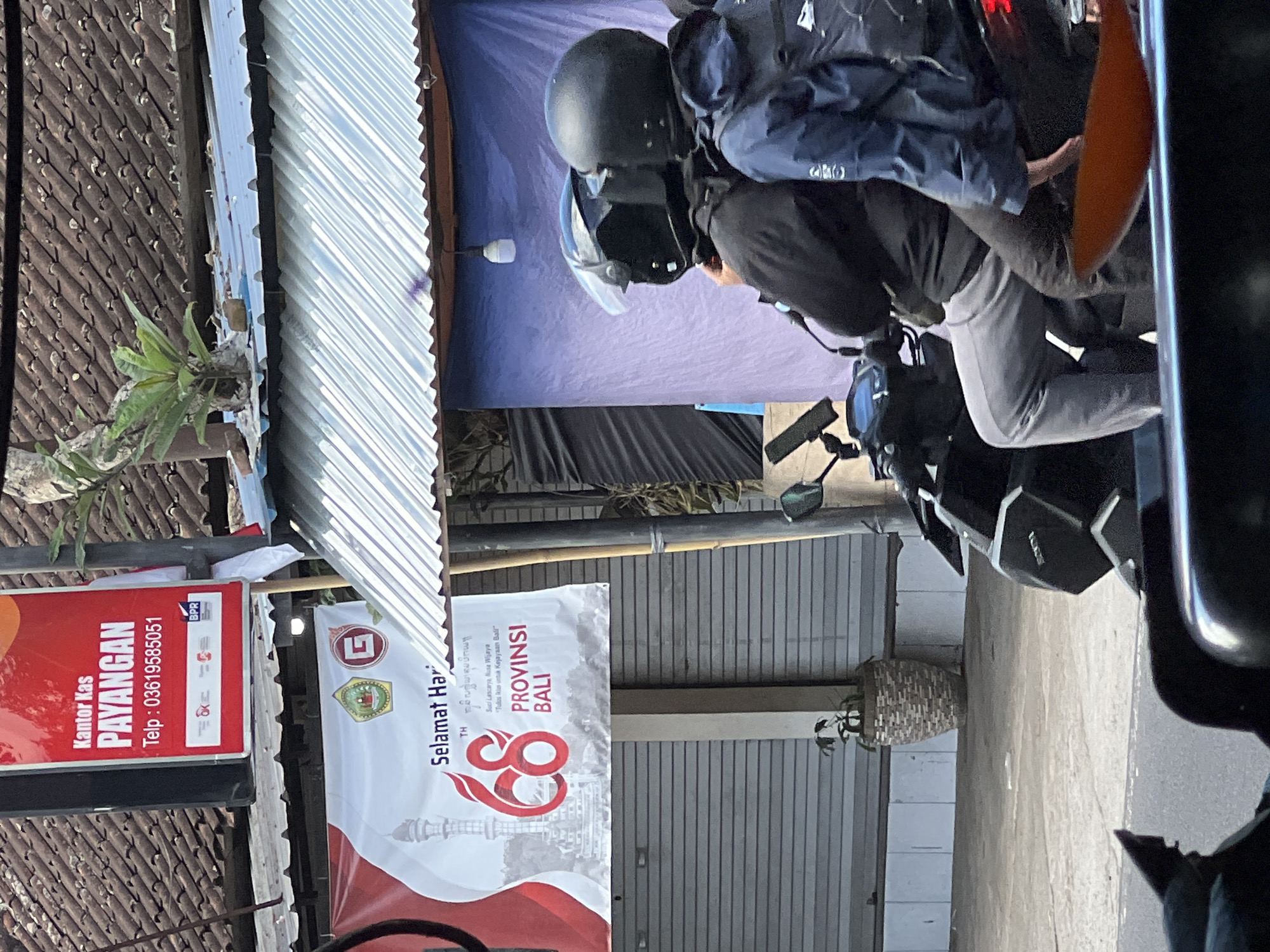 The image shows a street scene in Payangan, Indonesia. In the foreground, two people on a motorcycle are visible from the back, both wearing helmets. The person in the front is wearing a black jacket and grey pants, while the person in the back is wearing a blue jacket. The motorcycle is black. In the background, there is a banner celebrating "Selamat Hari... 68 PROVINSI BALI" (Happy Anniversary... 68 Bali Province). Above the banner, a red sign reads "Kantor Kas PAYANGAN" (Payangan Cash Office) with a phone number. The buildings have tiled roofs, and there is a plant in a decorative pot. The overall setting suggests a commercial or administrative area.