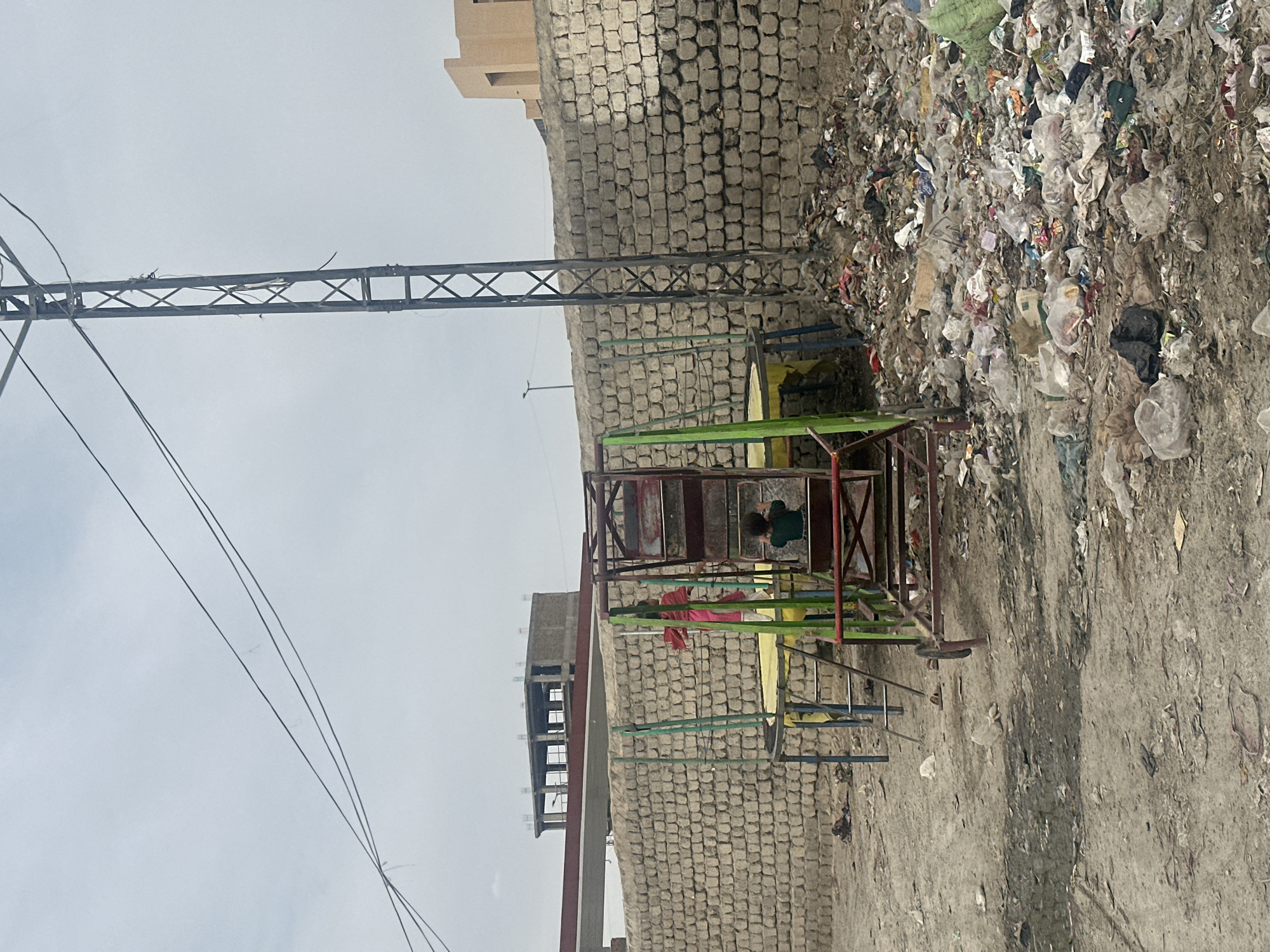 The image shows a scene in Quetta City Tehsil, Pakistan. A small, colorful, manually-operated merry-go-round is positioned near a brick wall. Debris and garbage are scattered around the base of the wall and on the ground. A child is visible on the merry-go-round. An electrical power pole is visible on the left side of the frame, with power lines extending across the sky. Buildings are seen in the background. The sky is overcast.