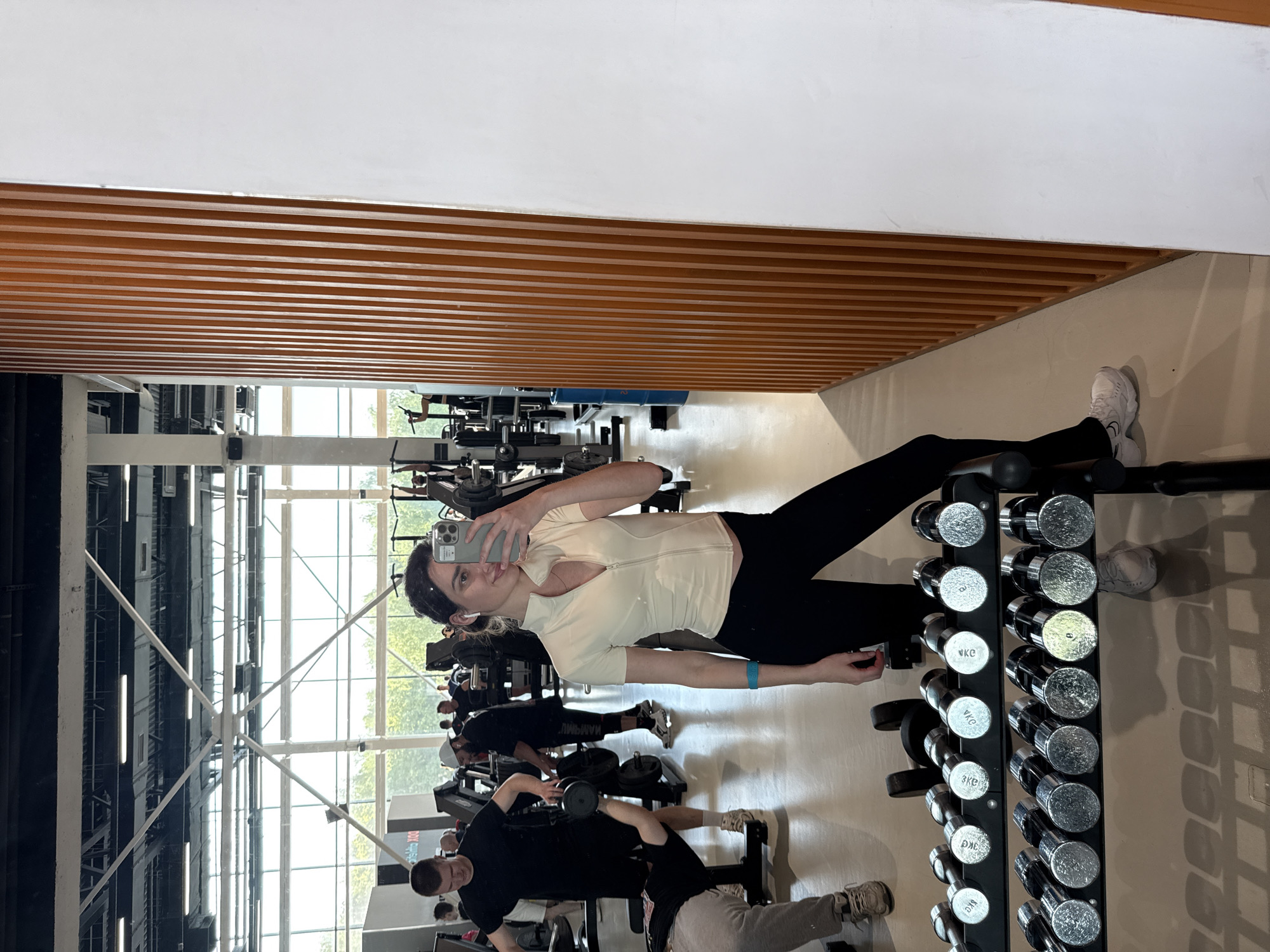 The image shows a woman taking a selfie in a gym. She is wearing a light-colored zip-up shirt and black leggings, and she is holding a phone up to her face. In the background, there are other people working out on various machines and using dumbbells. A rack of dumbbells is visible to the right of the woman, and the gym has large windows and high ceilings with visible support beams. The walls are a combination of light and dark colors, with a wooden slatted section near the ceiling.