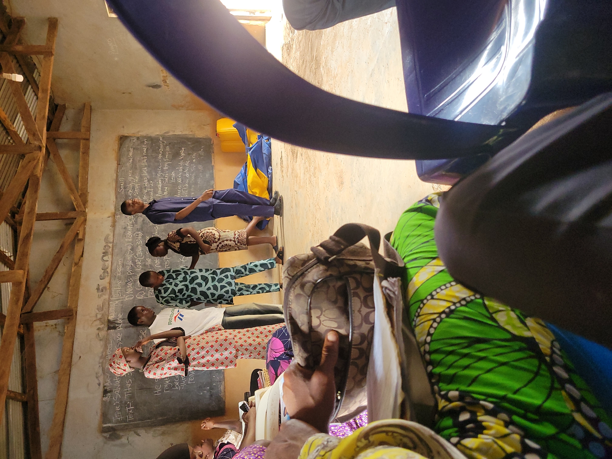 The image depicts a classroom in Unknown, Unknown. A row of students stands facing a chalkboard with handwritten text related to a Bible lesson, including topics such as salvation and grace. The chalkboard also includes sections written in Hausa. The classroom appears to be simply constructed with wooden support beams visible. A partially visible person in the foreground is holding a patterned handbag, and wearing a green and yellow patterned fabric.