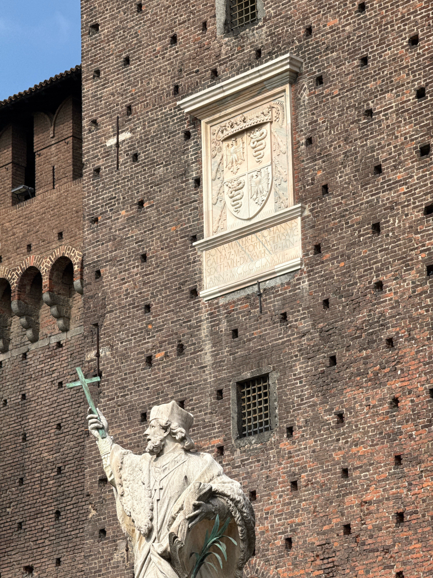The image shows a close-up of the brick facade of a castle, likely the Sforza Castle (Castello Sforzesco) in Milan, Italy. On the wall, there is a large, ornate stone crest with intricate carvings, possibly a coat of arms. Below the crest, there is an inscription in Latin. To the left of the crest, a section of the castle wall features arched windows. In the foreground, a statue of a robed figure, likely a religious or historical figure, stands holding a cross aloft. The figure is made of stone and is detailed, with a beard and a hat. The castle is constructed of dark red bricks and has small, square openings scattered across its surface. The sky is visible in the upper left corner, appearing blue and clear. The lighting suggests it is daytime.