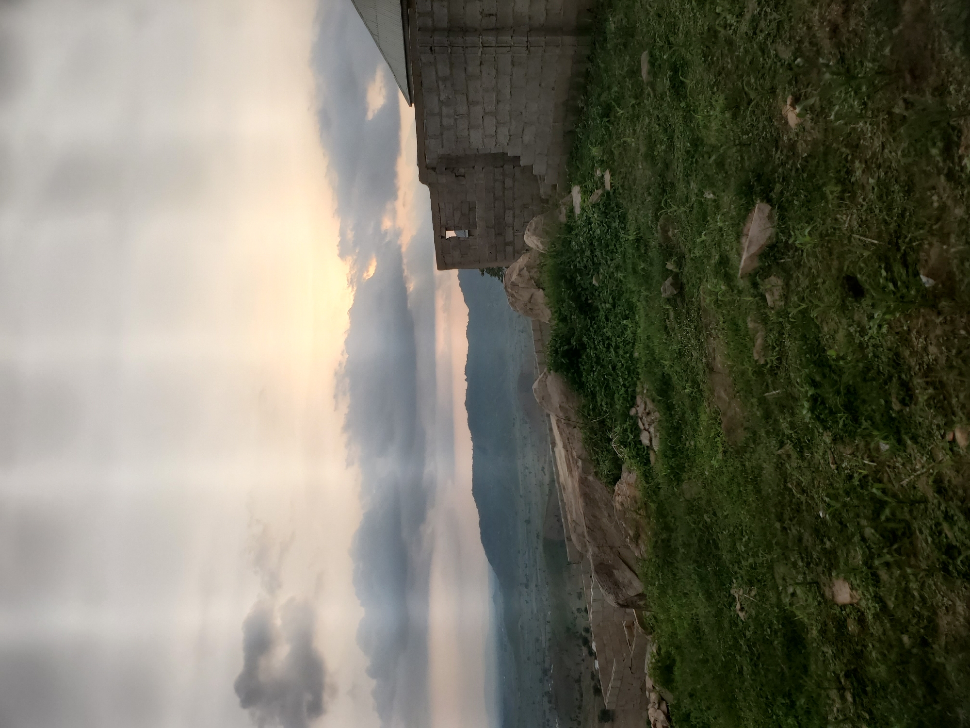 The image captures an outdoor scene with a cloudy sky at dusk. In the foreground, there is overgrown green grass and several large rocks. To the right, a partially constructed building made of cinder blocks is visible, with one section having a pitched roof. In the background, a lush green valley with rolling hills stretches out, partially obscured by atmospheric haze. The sky is a mix of dark and light clouds, with rays of sunlight breaking through from the upper right, illuminating parts of the sky and casting shadows. The overall impression is one of a rural or semi-developed landscape at the end of the day.