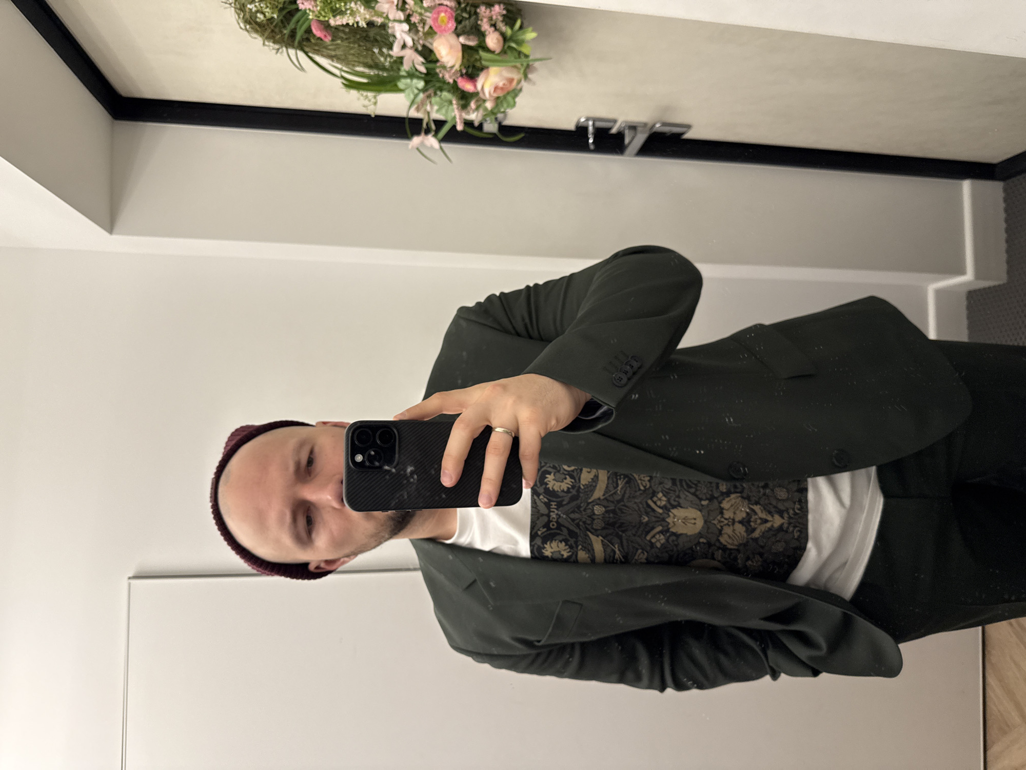 A man in a burgundy beanie and a dark green suit takes a selfie in a mirror. The mirror is decorated with a floral arrangement and what appears to be a faucet fixture. The location appears to be an indoor space, potentially a dressing room or bathroom, with a modern interior design. The exact location in Krasnoyarsk, Russia, cannot be determined from the image.