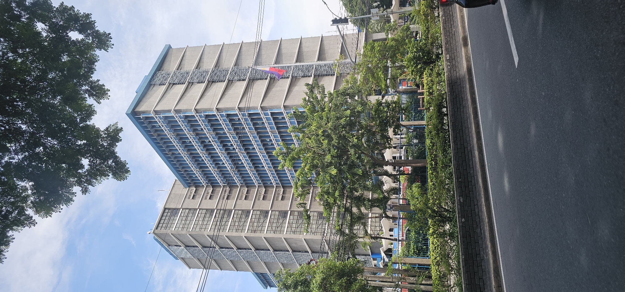 The image shows a tall, modern building with a gray facade and blue-tinted windows, located in Quezon City, Philippines. The building is multi-storied and appears to be an office building or government facility. A Philippine flag is visible on a flagpole in front of the building. The scene is an urban environment with trees and greenery in the foreground, suggesting a well-maintained area. A road runs in front of the building, with a black car partially visible on the right side of the frame. The sky is blue with some clouds. The architecture of the building suggests a contemporary design, possibly from the late 20th or early 21st century. The overall impression is that of a functional and important structure within a bustling city.