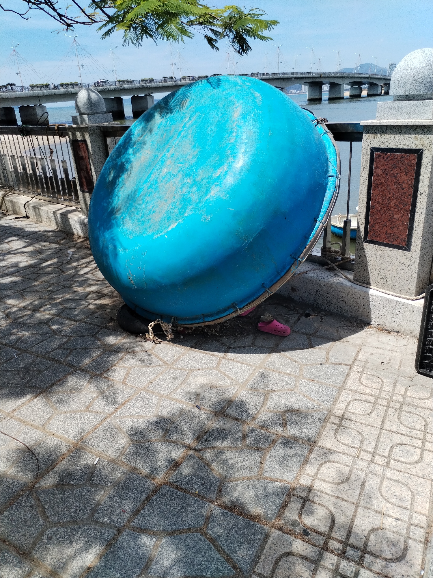 The image shows a large, upside-down blue coracle boat resting on a patterned stone pavement. To the left of the boat is a metal fence, and behind it, a body of water. A bridge spans the water in the background, with power lines and distant buildings visible. The scene is lit by daylight, casting shadows from nearby foliage onto the pavement. A pink sandal lies near the base of the boat. The location is the shoreline of the city of Nha Trang, Vietnam.